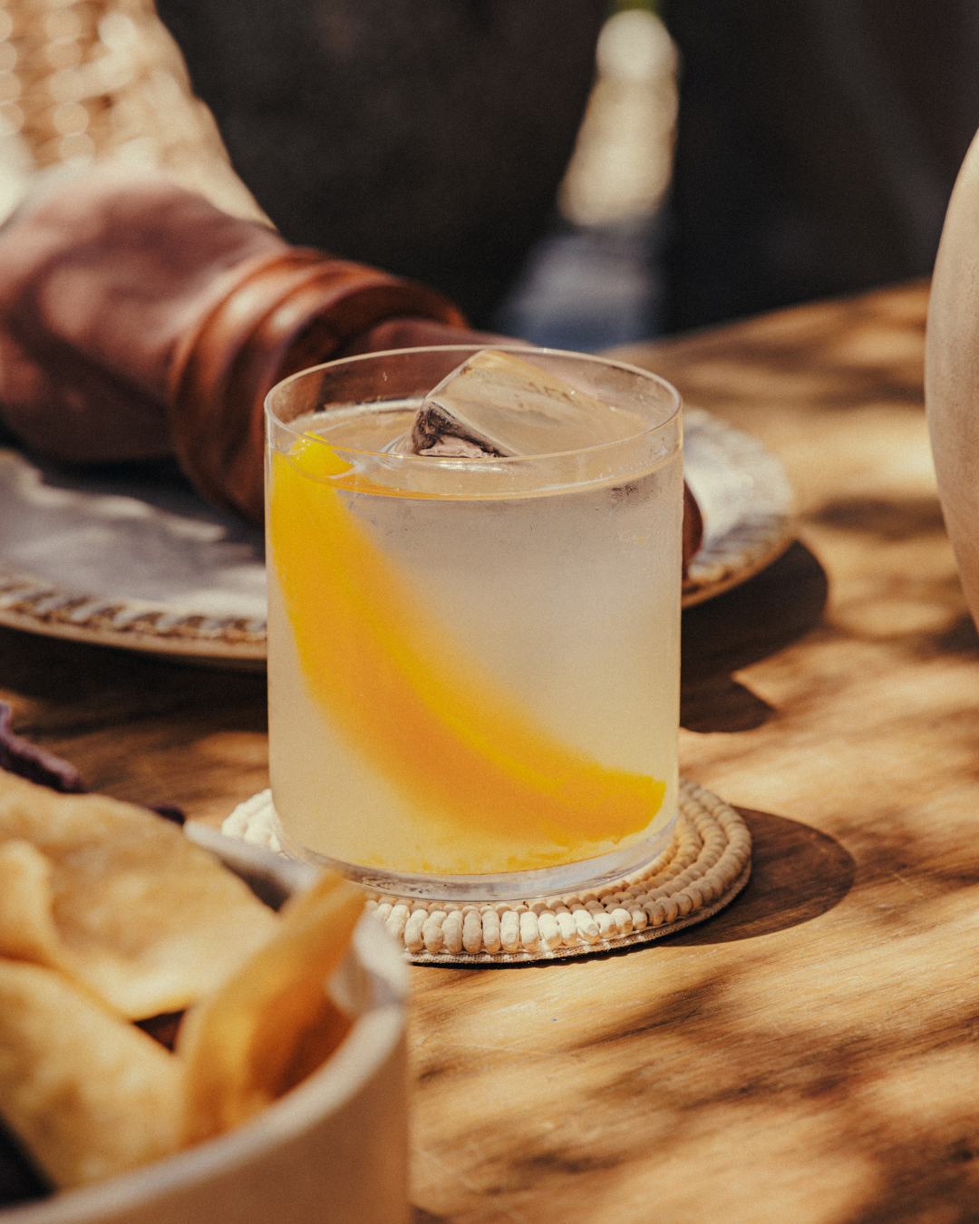 A bowl of multicolored plantain chips on a wooden table surrounded by two glasses of lemonade, bowls of soup, a basket, and plates with napkins.