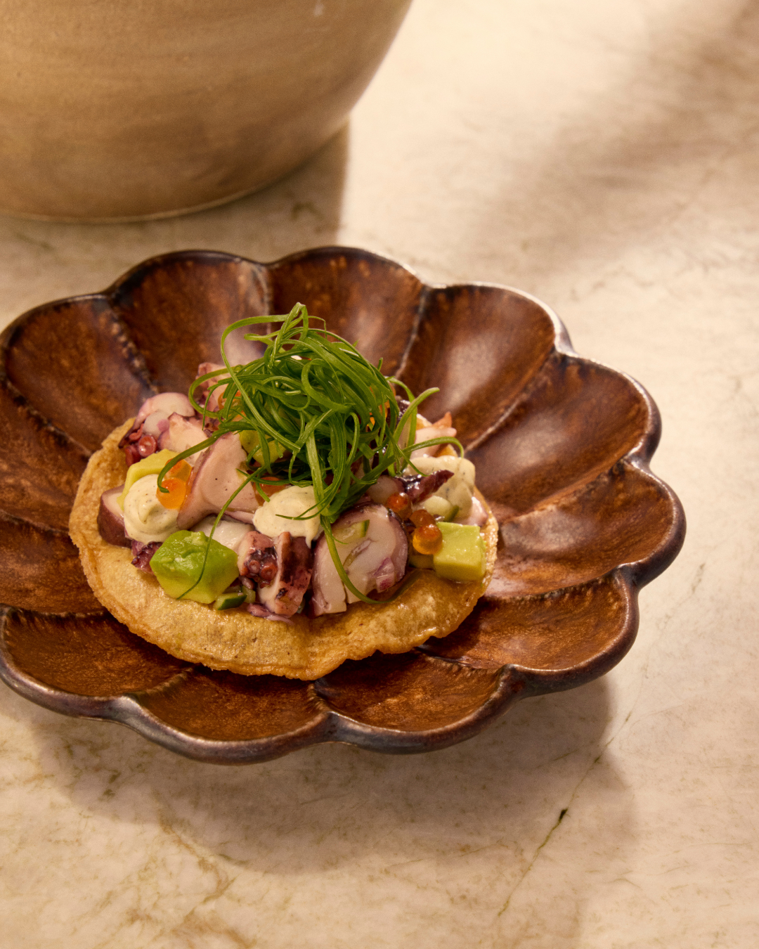A dessert featuring a chocolate cup with a scoop of ice cream, topped with a yellow fruit compote, a mint leaf, and small black berries, served on a speckled beige plate with crushed chocolate and cocoa powder.