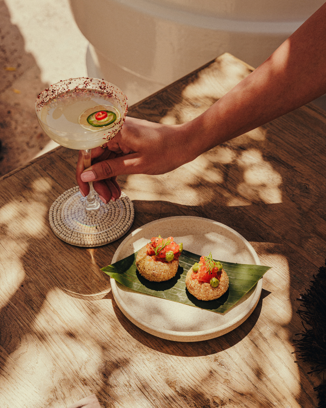 A person holding a cocktail glass with a lime and red chili pepper garnish, next to a plate of two sushi rolls on a banana leaf.