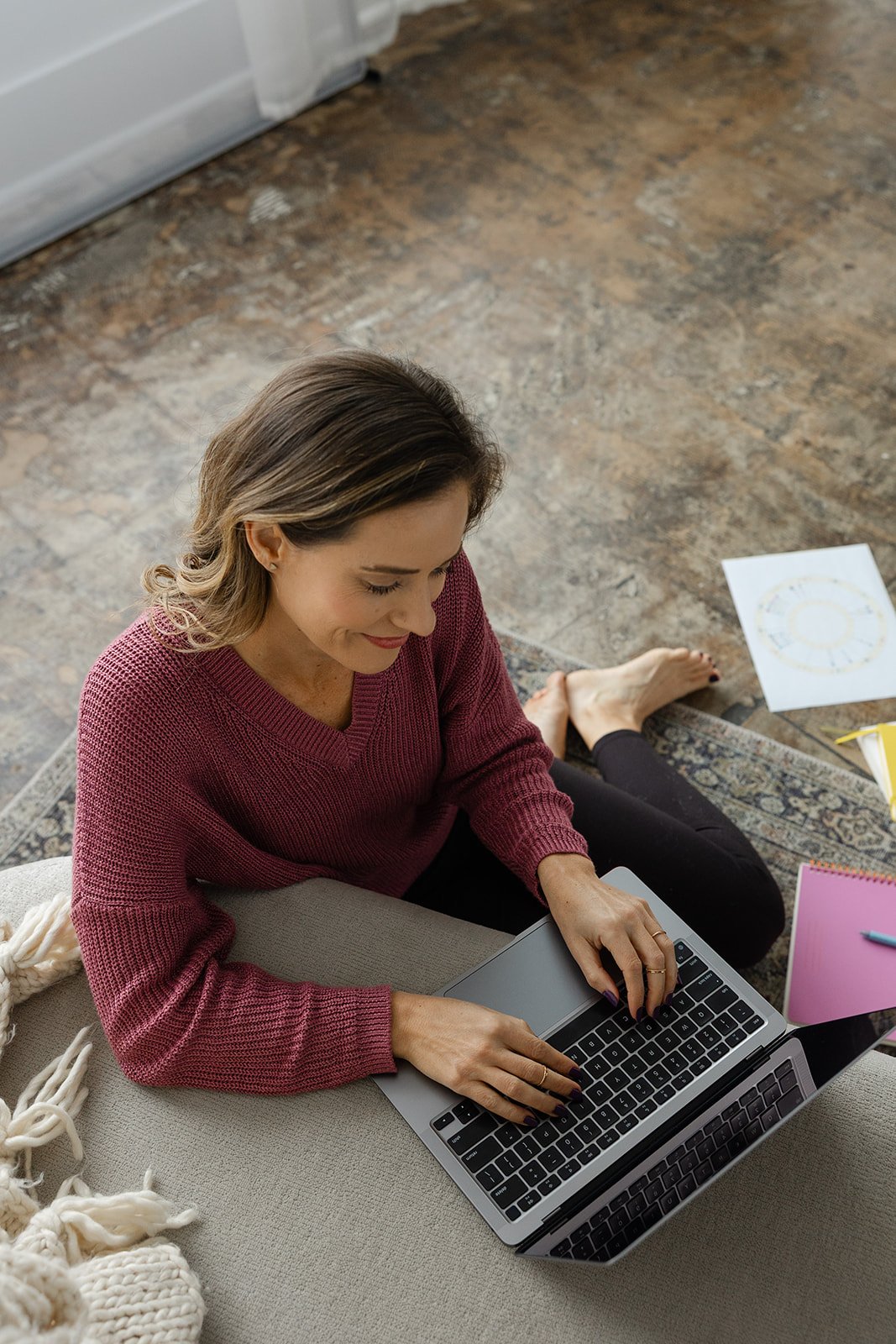 A woman in a red sweater sitting on a beige sofa, working on a laptop in a room with wooden flooring, with papers and notebooks nearby.