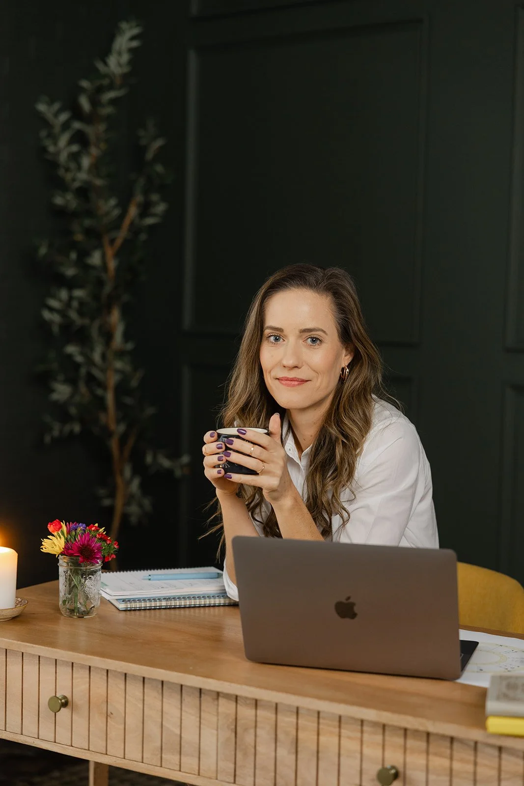 A woman with long wavy brown hair and wearing a white shirt is sitting at a wooden desk, holding a black coffee mug, and looking at the camera. There is a laptop, a notepad, a pen, a small vase with colorful flowers, a lit candle, and some magazines on the desk. The background is dark with a tall plant and a dark wall.