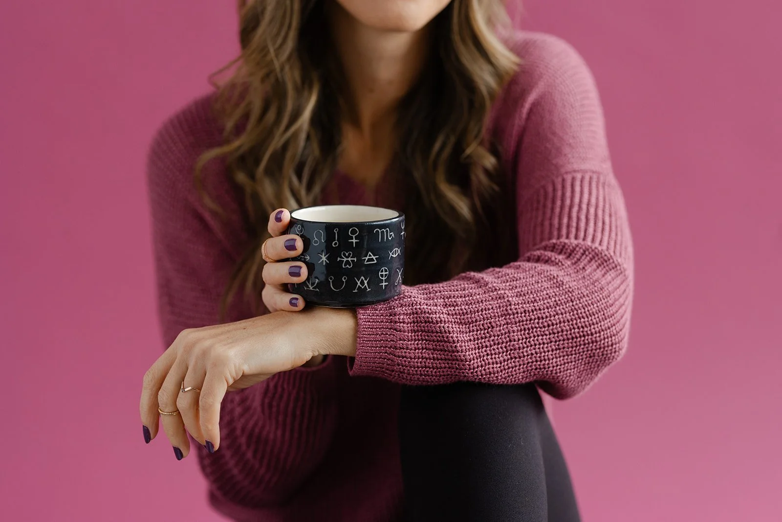 Woman with wavy brown hair wearing a pink sweater, holding a black mug with astrological symbols on it against a pink background.