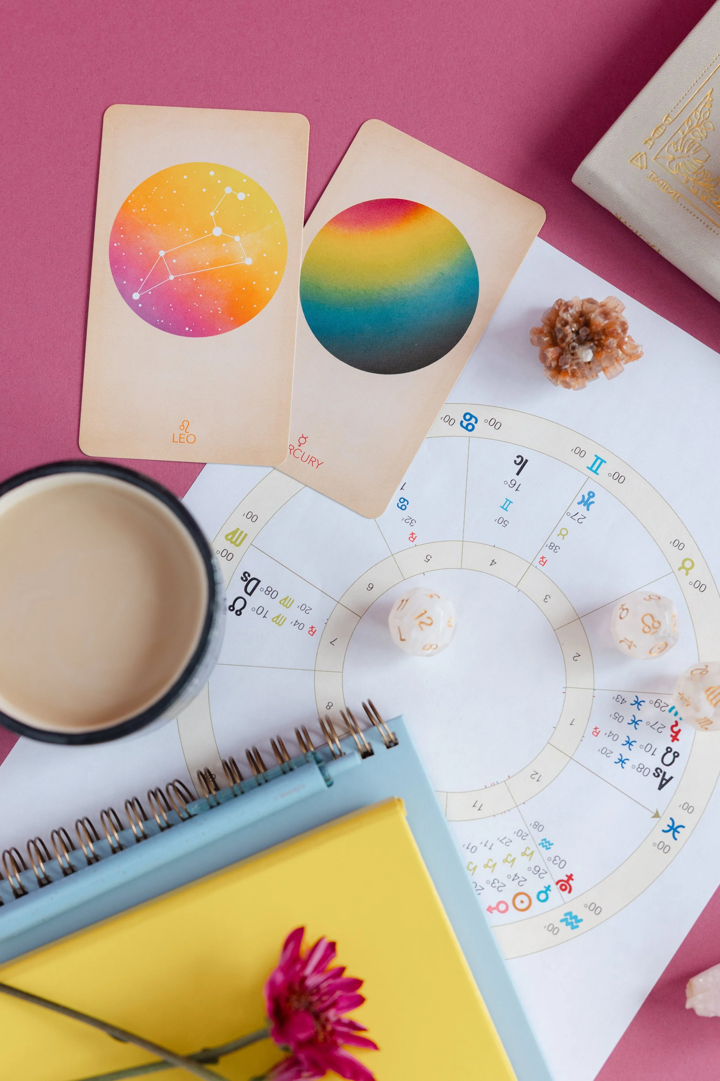 A flat lay of astrology and zodiac-themed items, including two oracle cards with colorful circular designs labeled Leo and Pluto, a spiral-bound notebook with a light blue cover and a yellow cover with a pink flower, a paper with astrological charts, white dice with orange numbers, a cup of coffee or tea, a small orange mineral, and a white box with gold accents on a pink background.