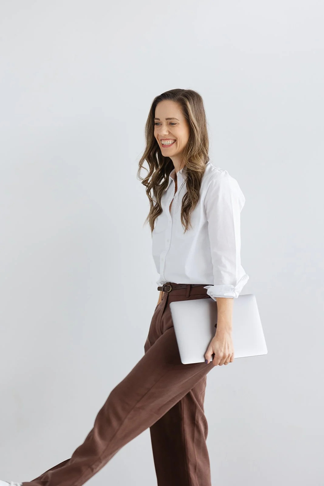 A woman smiling and walking, holding a closed laptop in her right hand.