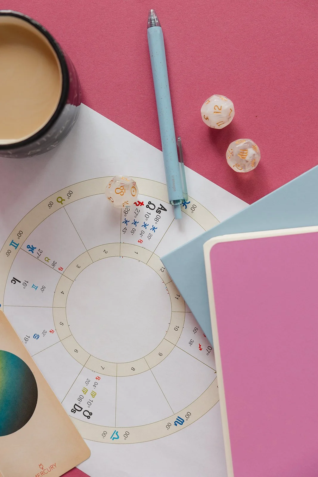 An astrology chart, a blue pen, a white and pink dice with numbers, a cup of coffee, and a booklet on a pink and white background.