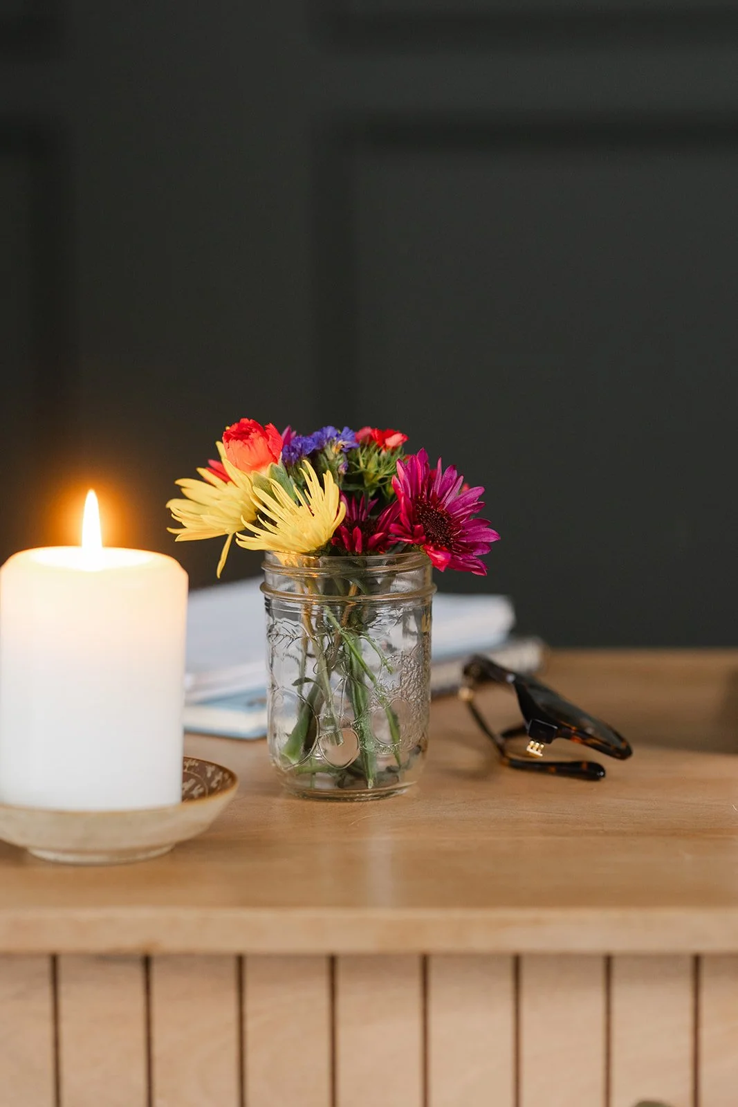 A small bouquet of colorful flowers in a glass jar, a lit white candle in a holder, a pair of glasses, and some papers on a wooden surface with a dark background.
