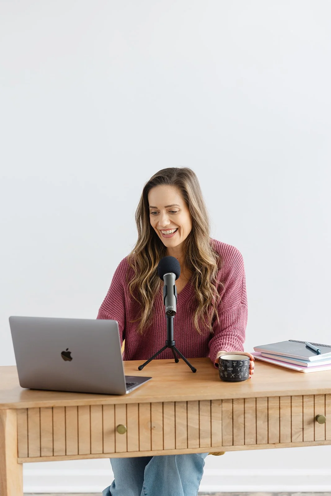 Woman in a pink sweater speaking into a microphone, sitting at a wooden desk with a laptop, notebooks, and a coffee mug.