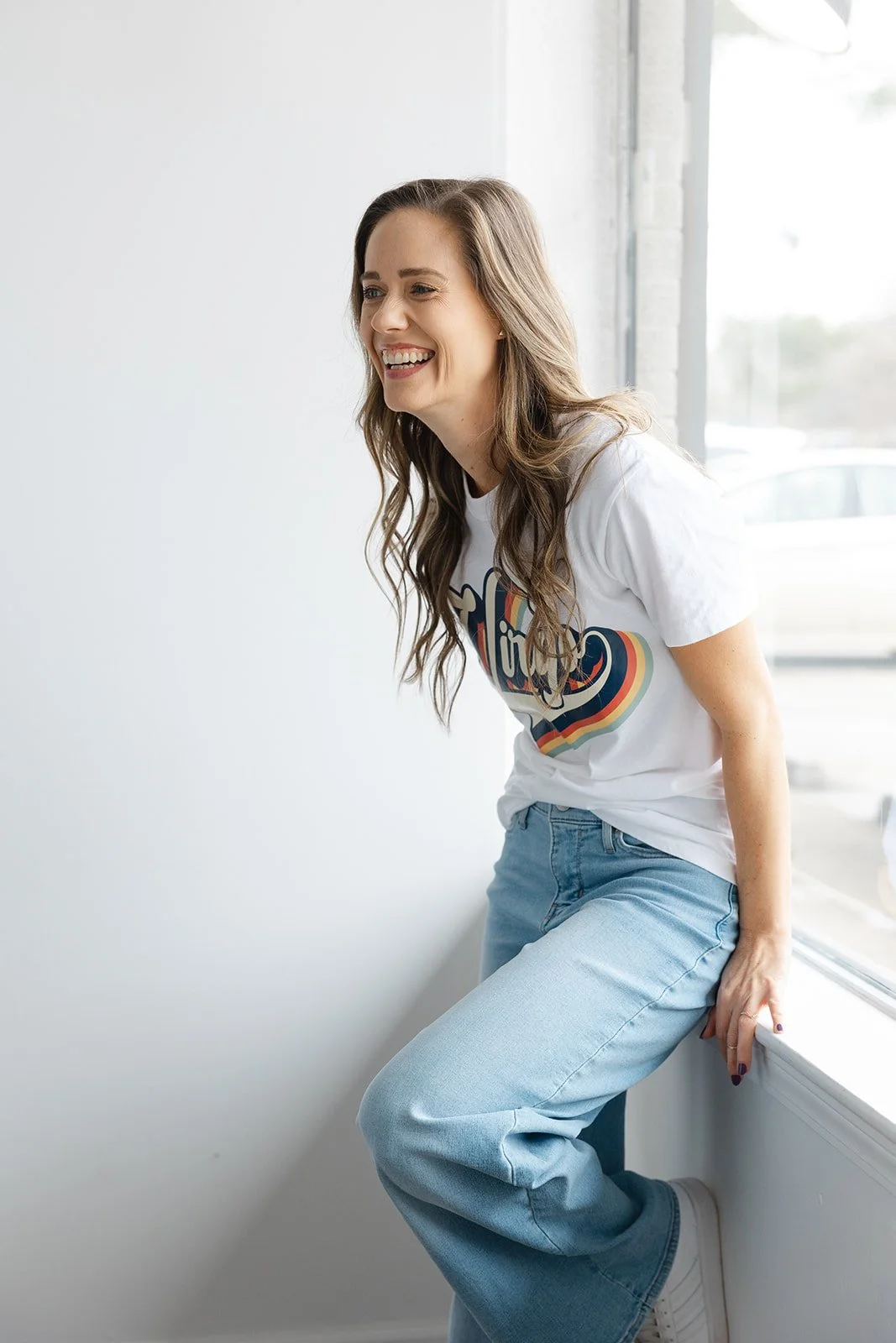A woman with long brown hair wearing a white t-shirt and blue jeans sitting by a window, smiling and leaning against the windowsill.