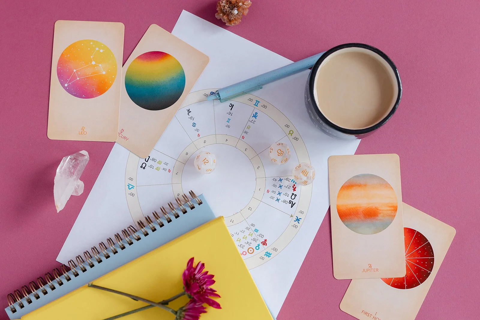 A pink workspace with astrology cards, a cup of coffee, a clear crystal, and a colorful astrology chart with a notebook and pink flower.
