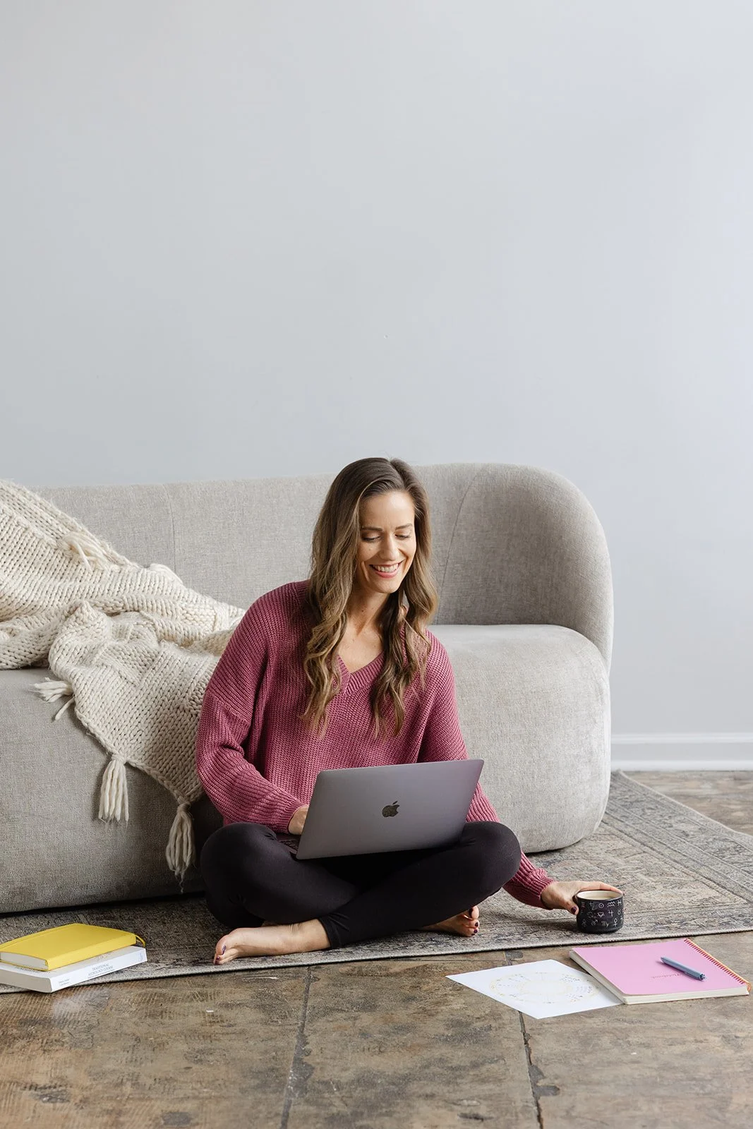A woman sitting cross-legged on a rug in front of a sofa, working on a laptop. She is holding a mug, with notebooks, papers, and a pen on the floor around her.