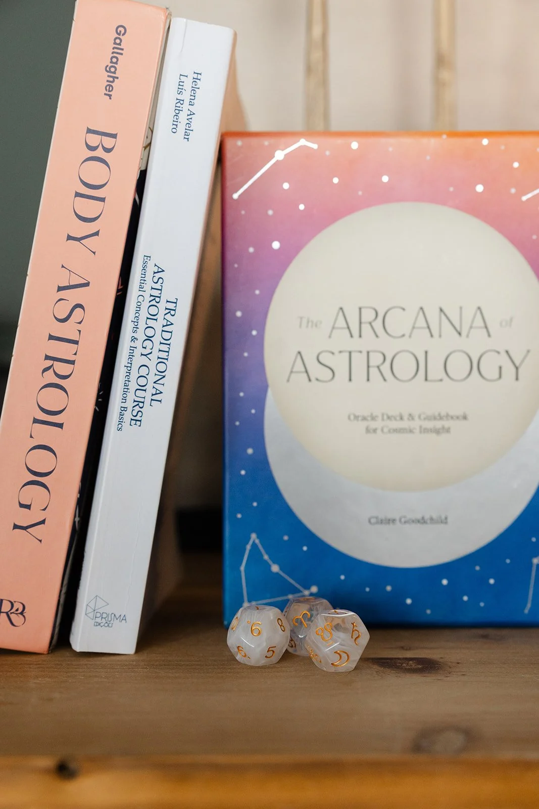 A stack of three books about astrology, including 'The Arcana of Astrology' in the foreground, with a set of three translucent astrology-themed dice on a wooden surface in front of the books.
