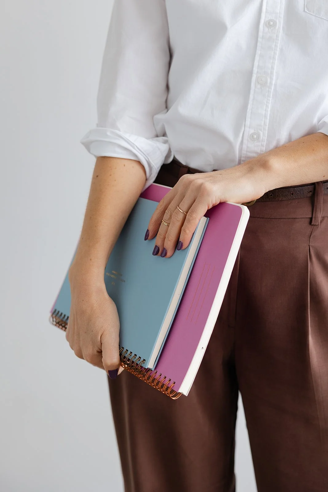 A person in a white shirt holding three notebooks, two spiral-bound, one pink and one blue, with brown pants.