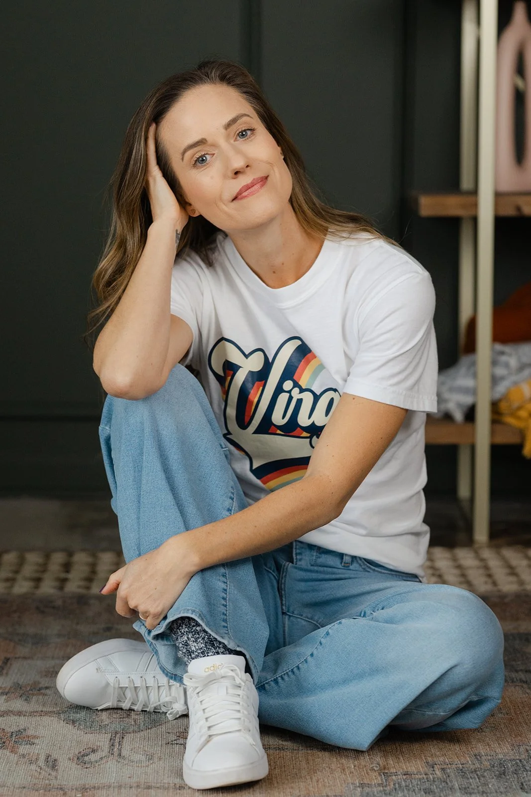 A woman with light brown hair sitting on the floor, wearing a white t-shirt with a colorful retro design, blue jeans, and white sneakers, resting her head on her hand and smiling slightly.
