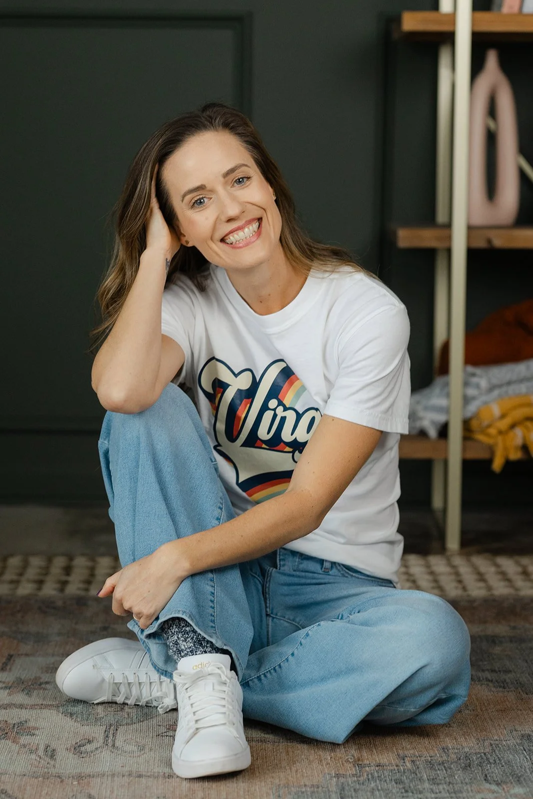 A smiling woman with long brown hair sitting on the floor, wearing a white T-shirt with colorful text, light blue jeans, and white sneakers, in a room with a dark green wall and wooden shelves.