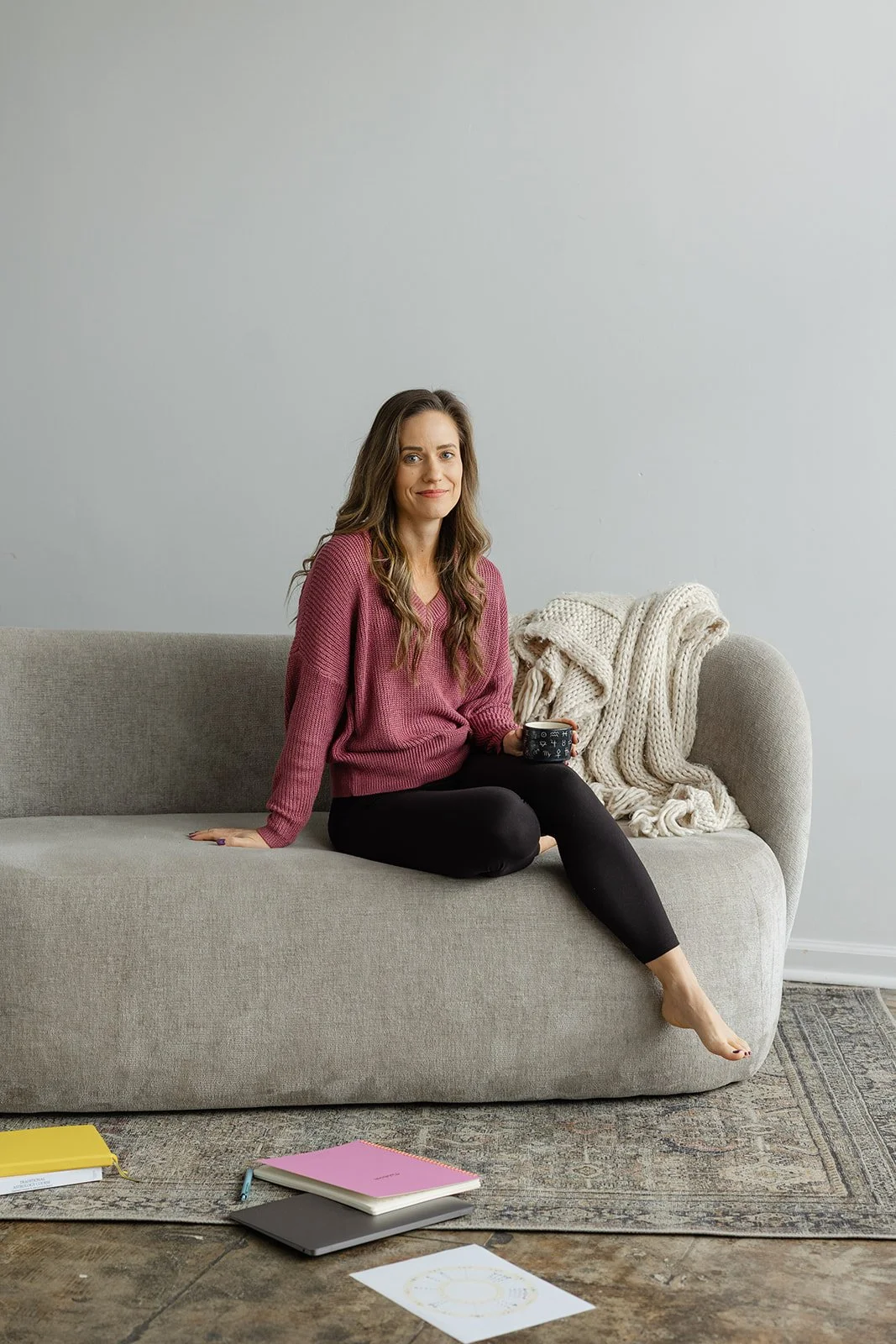 A woman with long brown hair sitting on a beige sofa, holding a mug, with a cream-colored chunky knit blanket beside her, in a room with a plain white wall.