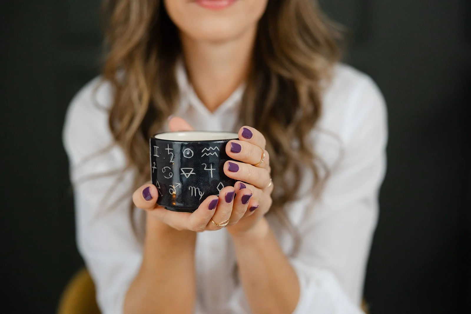 A woman with wavy brown hair and purple nail polish holds a black mug with astrological symbols on it, wearing a white shirt.