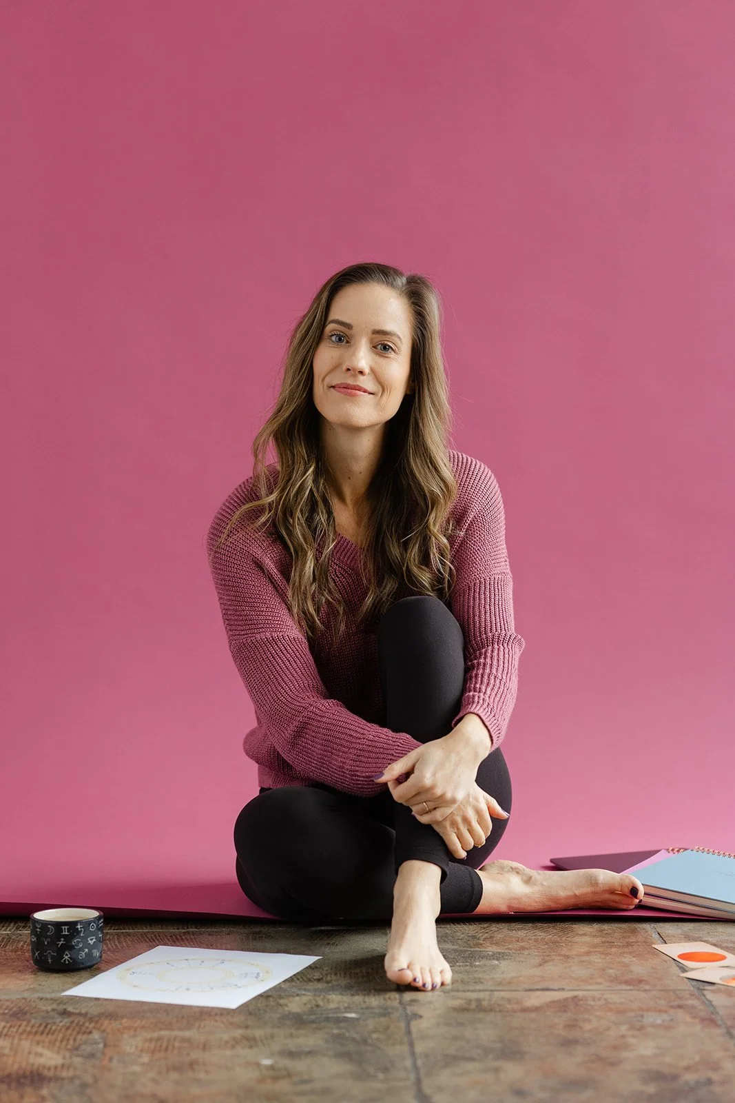 A woman with long wavy brown hair sits barefoot on a wooden floor against a pink background, wearing a pink sweater and black leggings, smiling at the camera. Items on the floor include a black mug with white symbols, a sketch, and colorful notebooks.