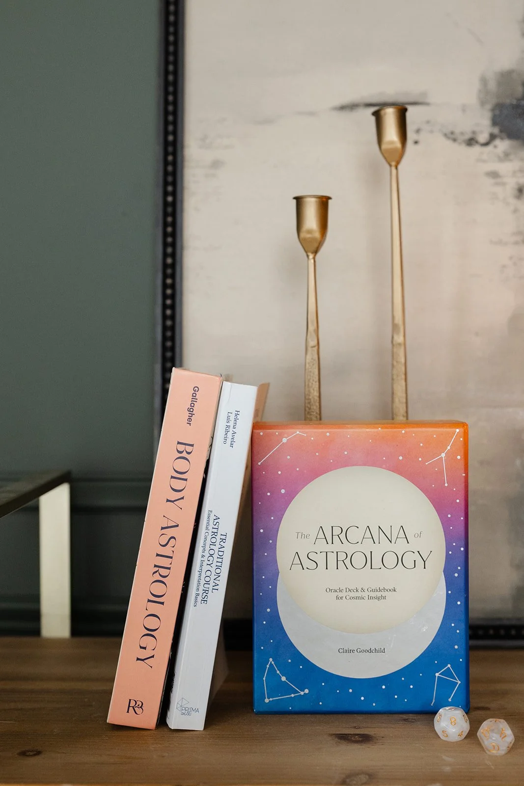 Books on astrology and body astrology, two white dice with gold symbols, and two tall gold candle holders on a wooden surface.