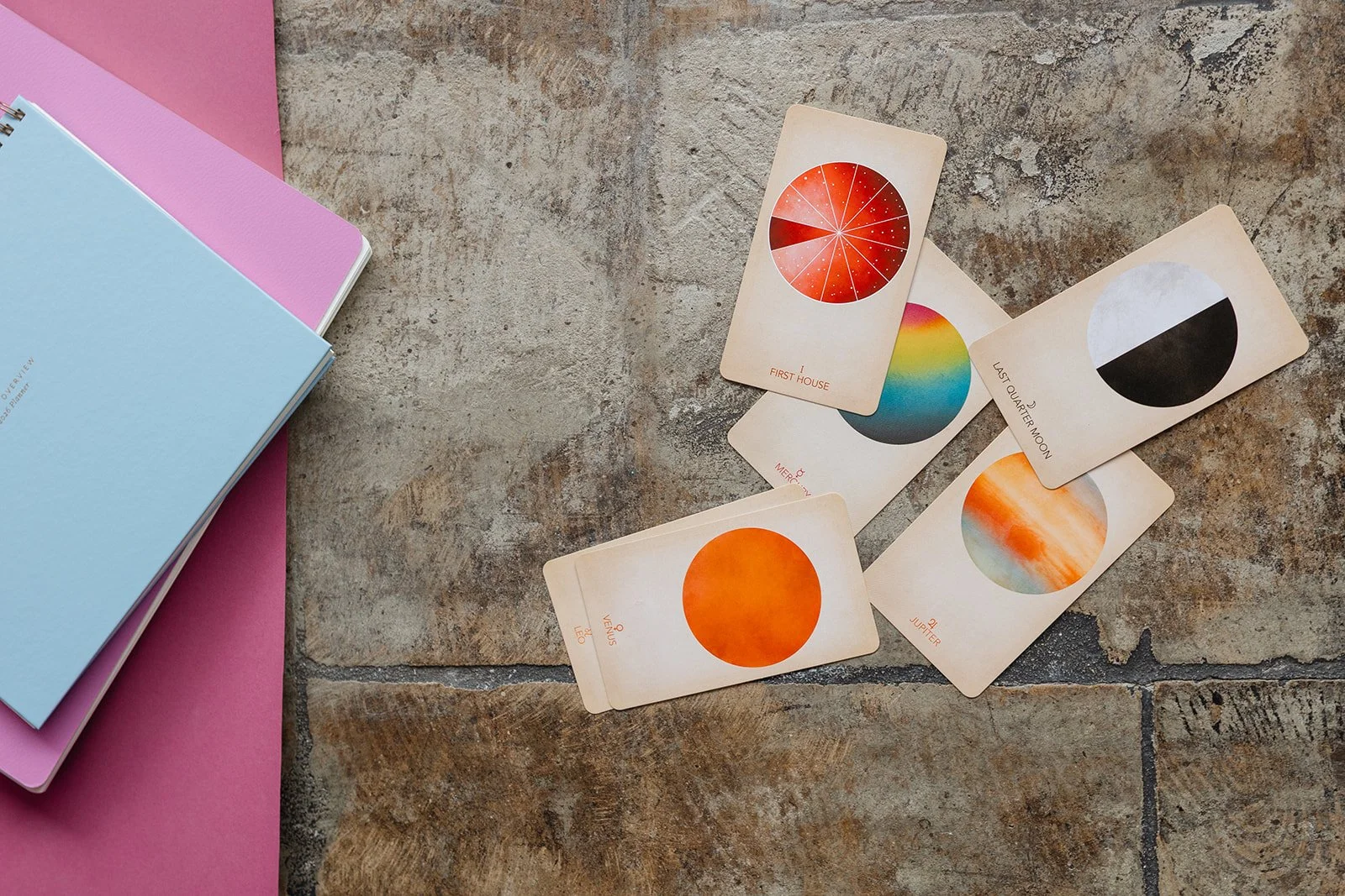 Colorful constellation or planet-themed cards spread out on a rustic wooden surface with some pink and blue books or notebooks nearby.
