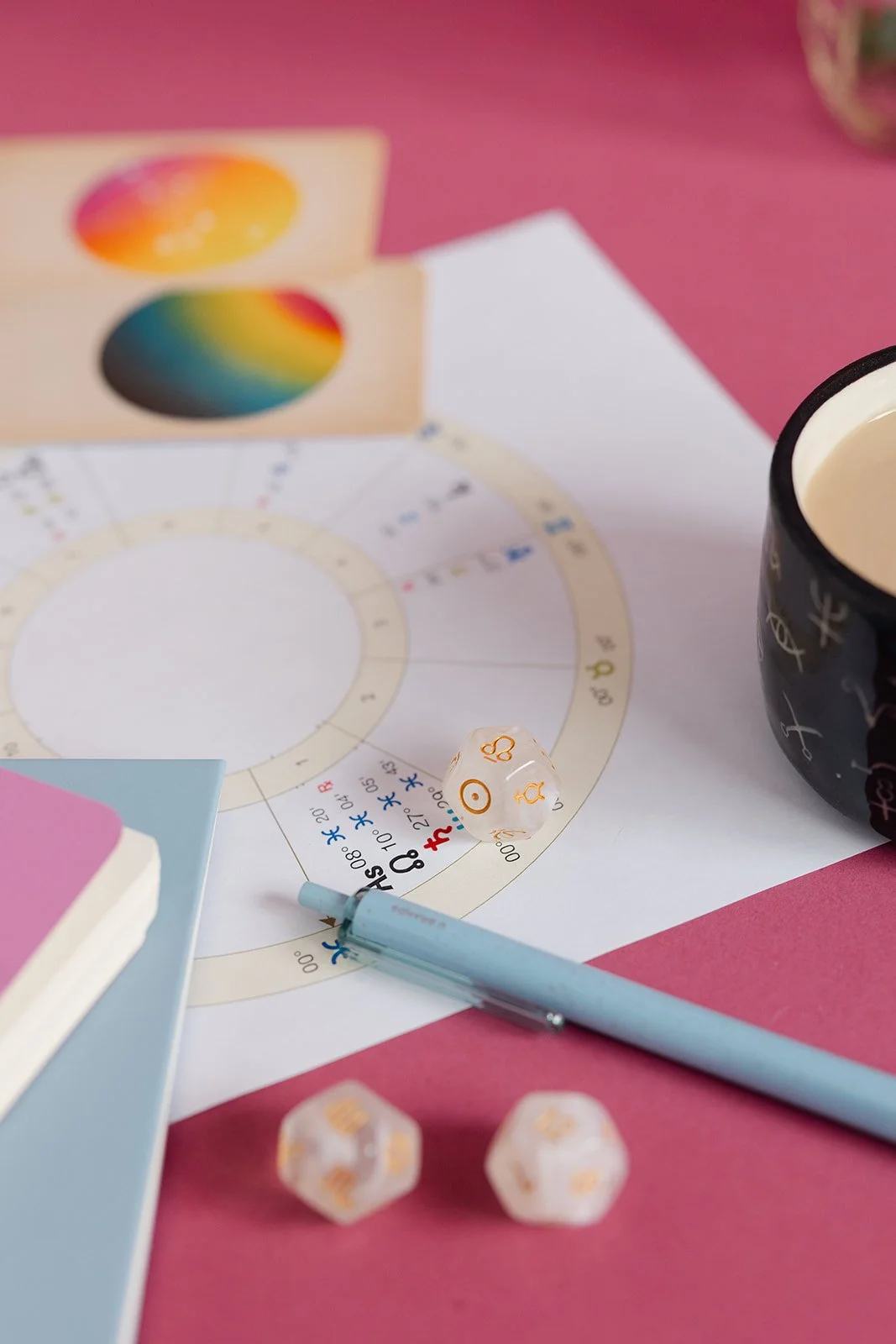 Board game setup with a color wheel, two white dice with orange symbols, a light blue pen, a pink and white book, a black mug with writing, on a pink surface.