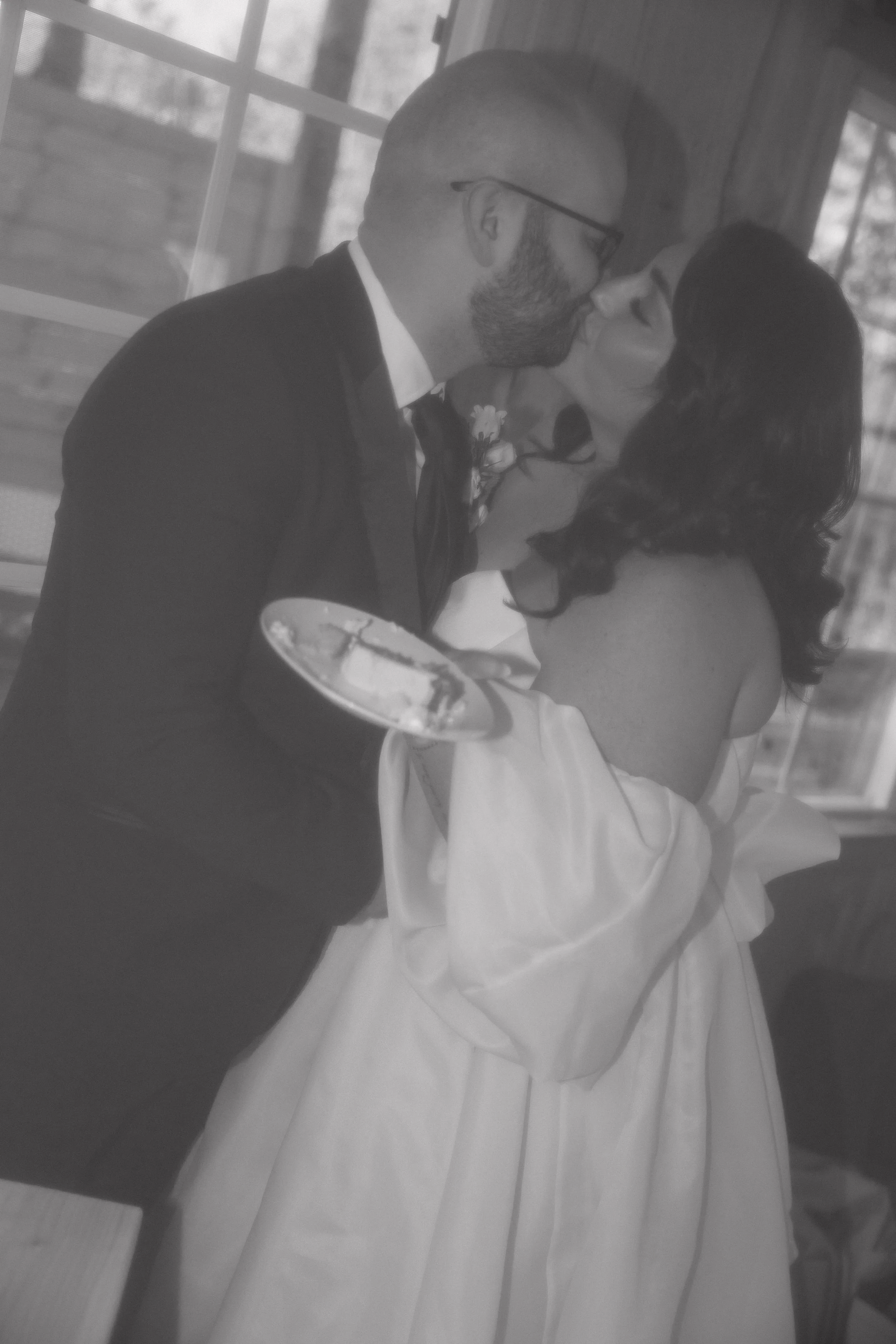 A black-and-white photo of a couple sharing a kiss during their wedding, with the groom in a suit and the bride in a strapless dress, holding a cake server.
