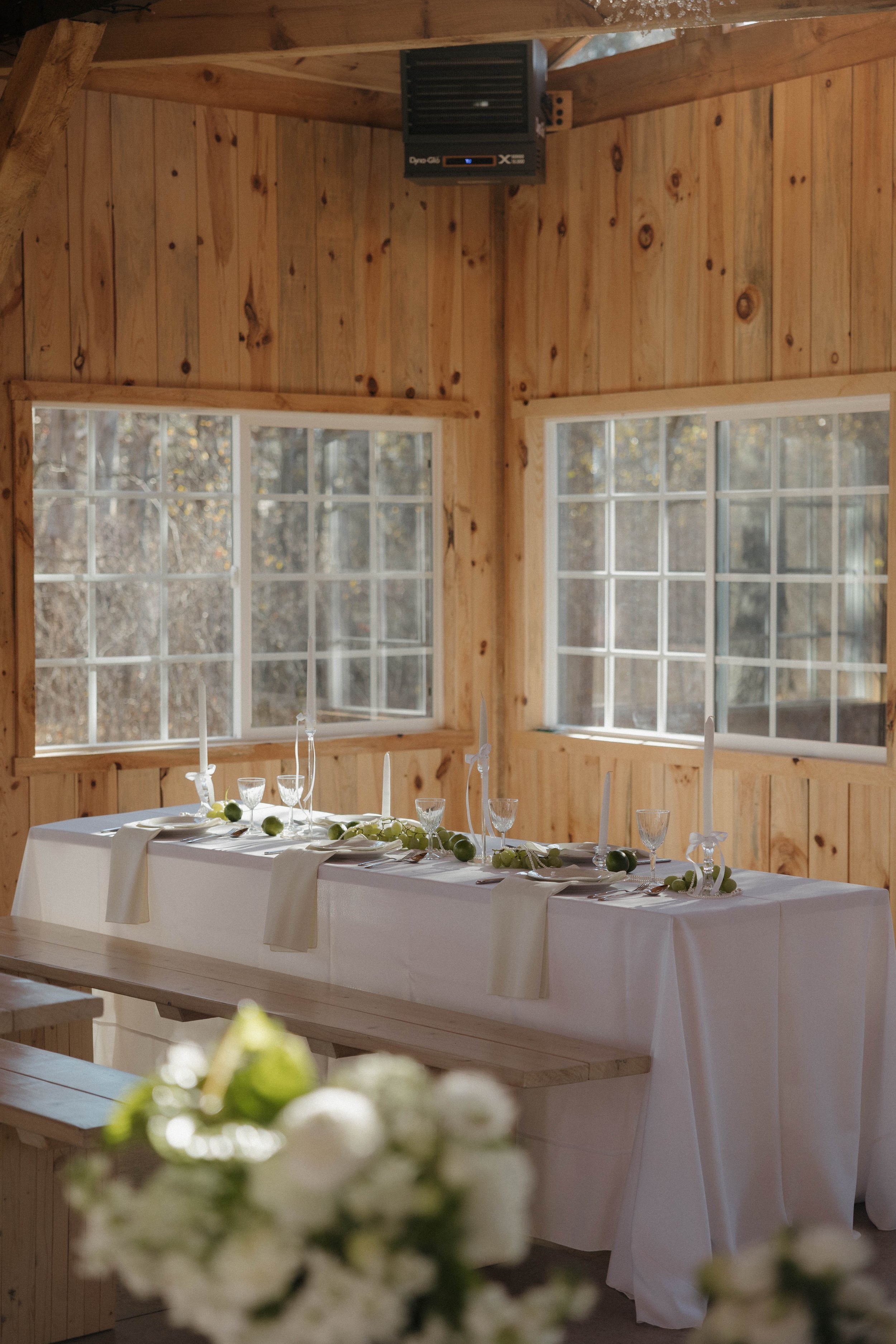 Decorated wedding reception table in a rustic wooden room with large windows and a bouquet of white flowers in the foreground.