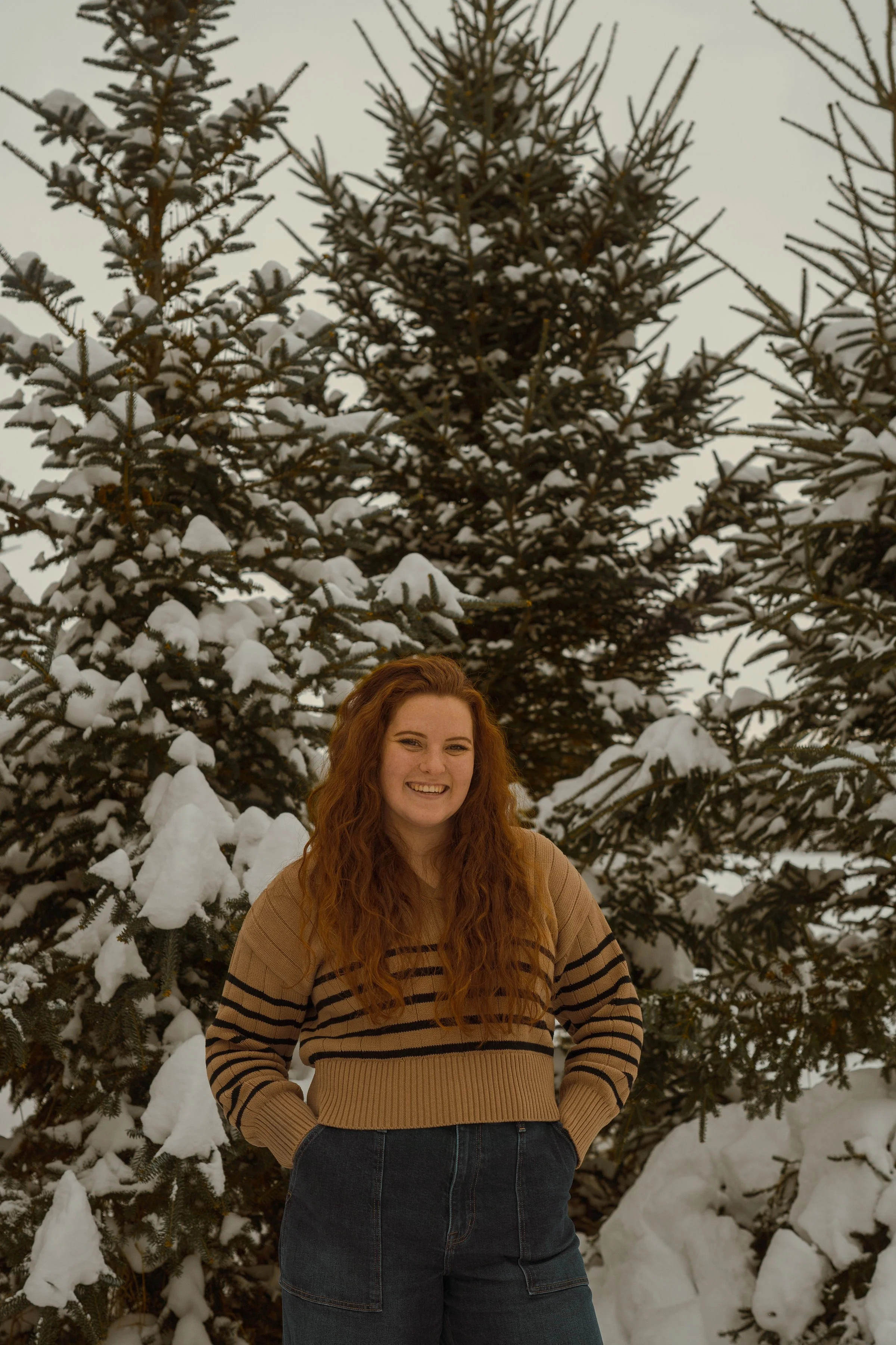 A young woman with long, curly red hair smiling and standing outdoors in front of snow-covered evergreen trees on a winter day, wearing a tan and black striped sweater and dark jeans.