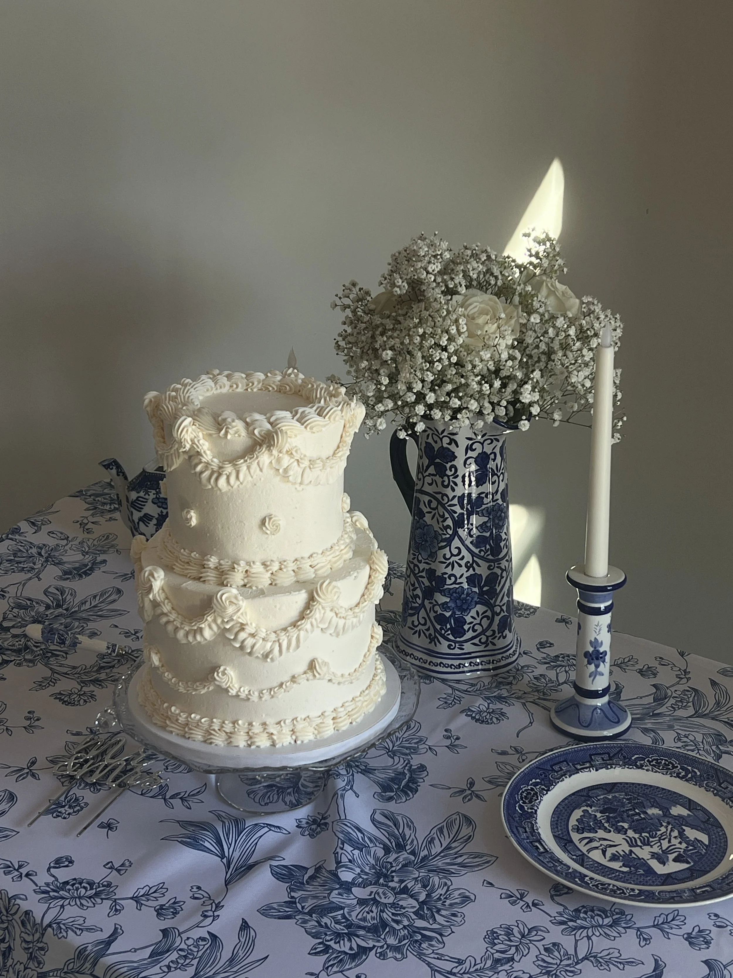 A three-tiered white wedding cake decorated with piped frosting, a vase of white flowers, a lit candle, and a blue and white floral-patterned plate on a matching tablecloth.