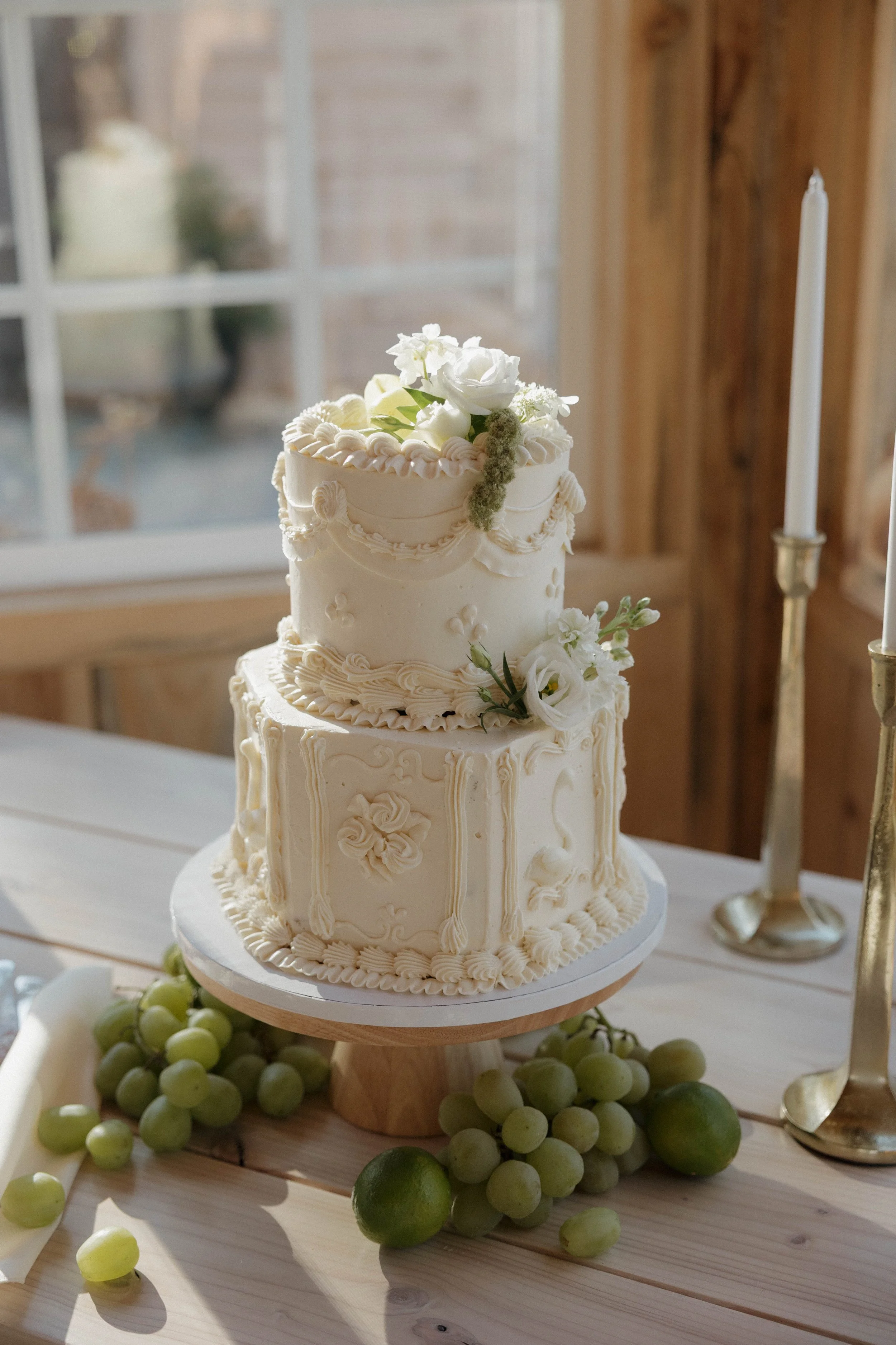 Two-tiered white wedding cake decorated with flowers and white frosting, placed on a wooden table with green grapes and two gold candlesticks nearby.
