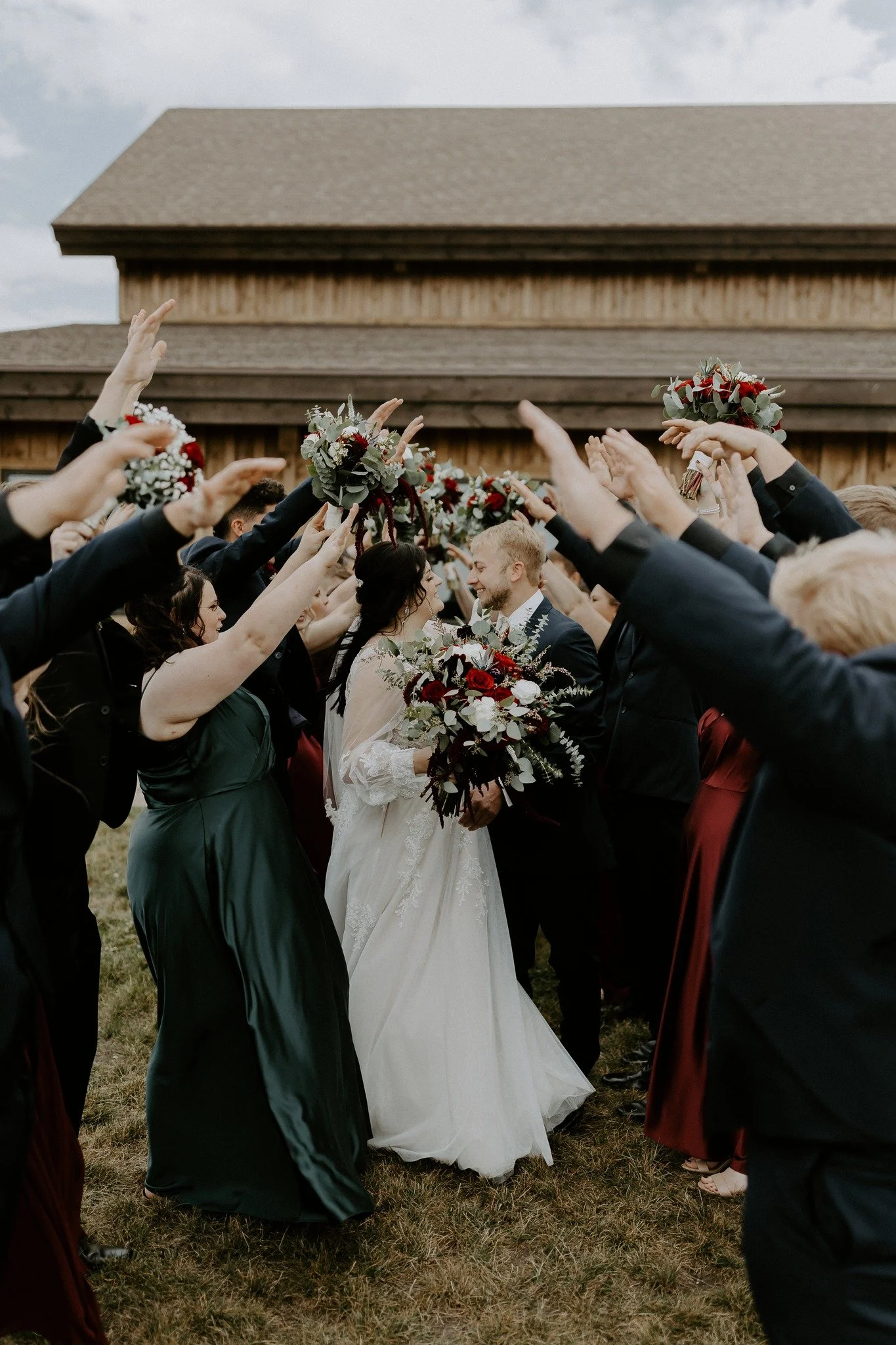 A bride and groom walking through an arch formed by wedding guests holding bouquets and raising their hands, outside in front of a rustic wooden building.