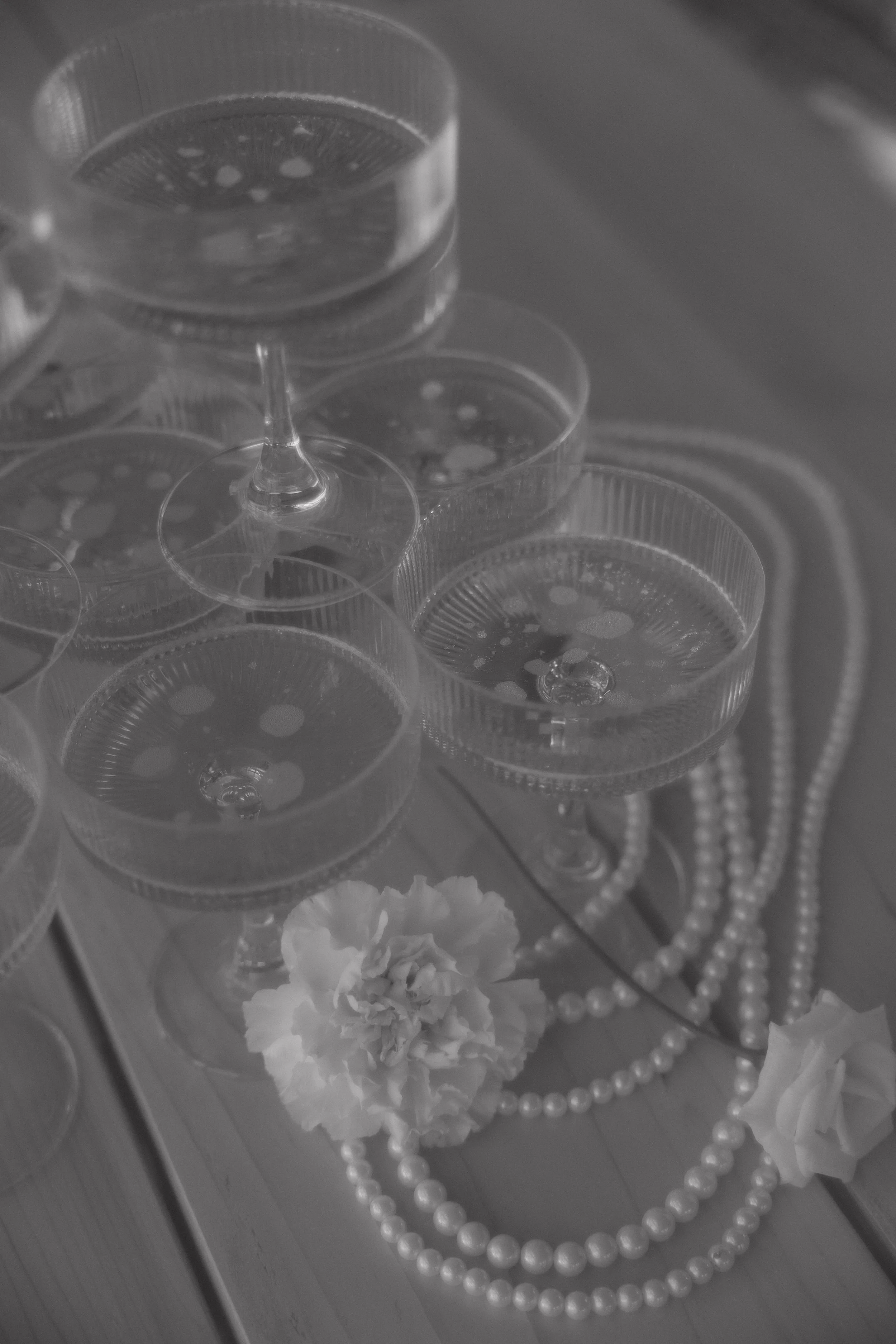 A stack of clear cocktail glasses with bubbles inside, a string of pearls, and artificial flowers on a wooden surface.