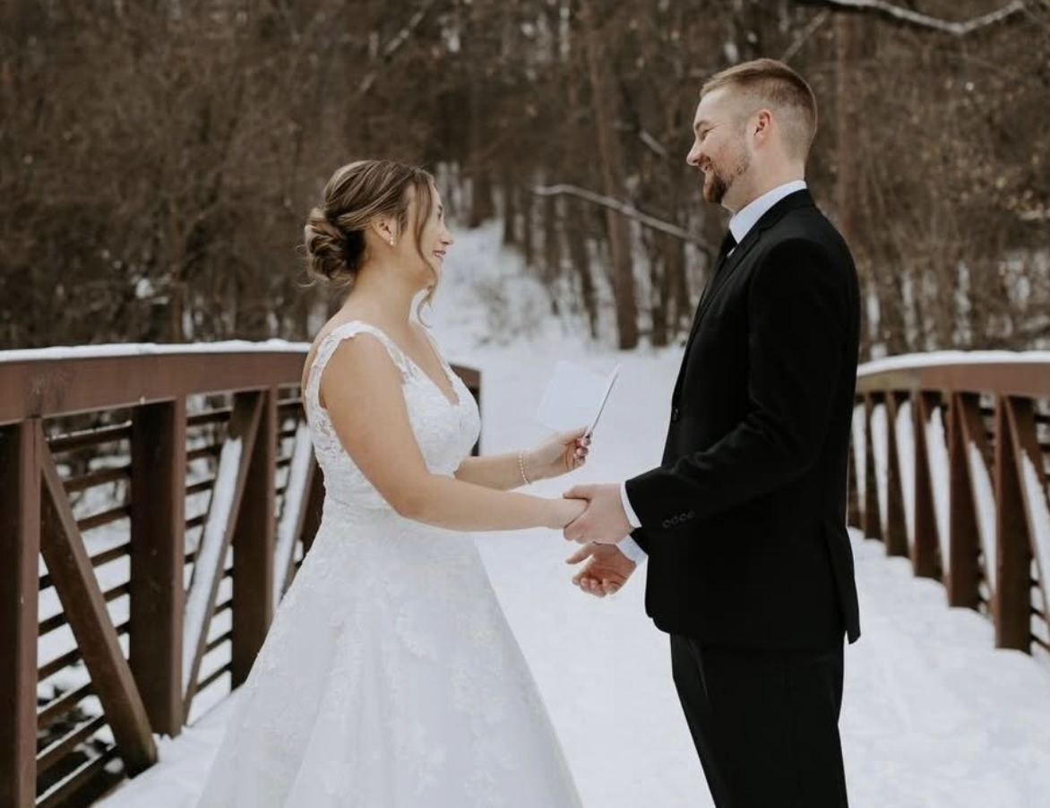 A bride and groom exchange vows outdoors on a snowy bridge during winter, holding hands and smiling at each other.