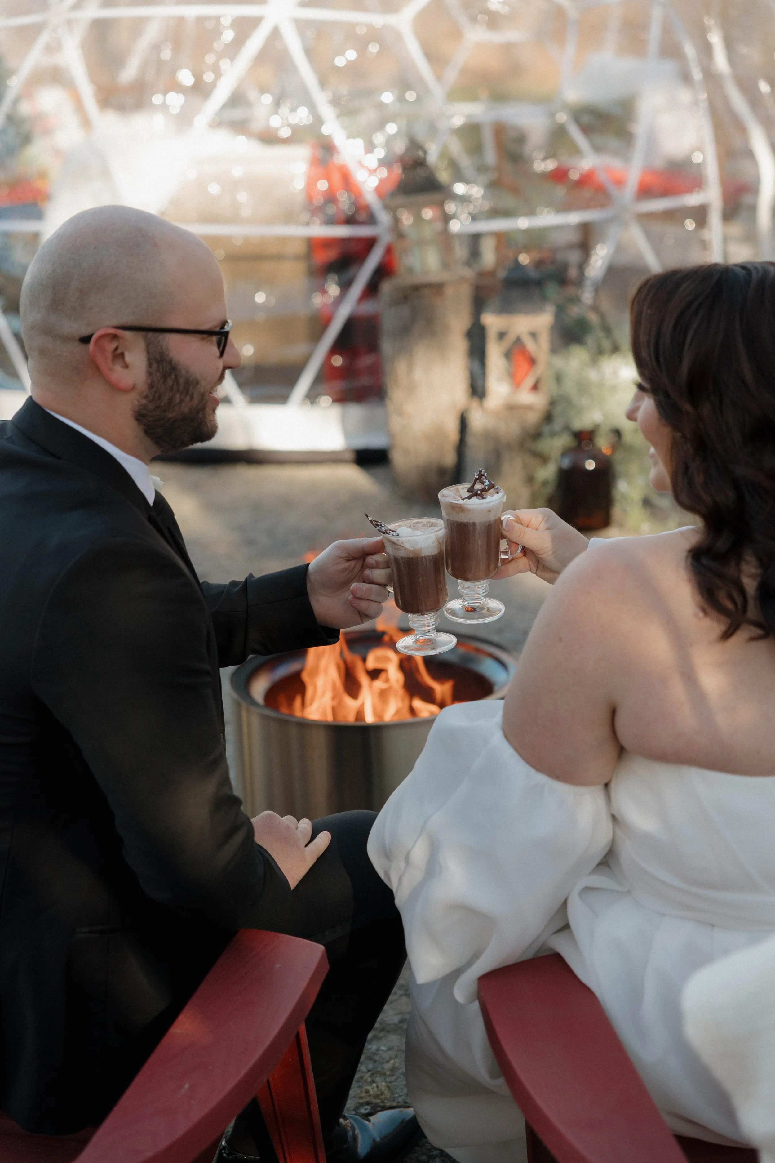 A couple dressed formally, sitting outdoors next to a fire pit, sharing a toast with chocolate drinks garnished with chocolate shavings, during sunset or evening.