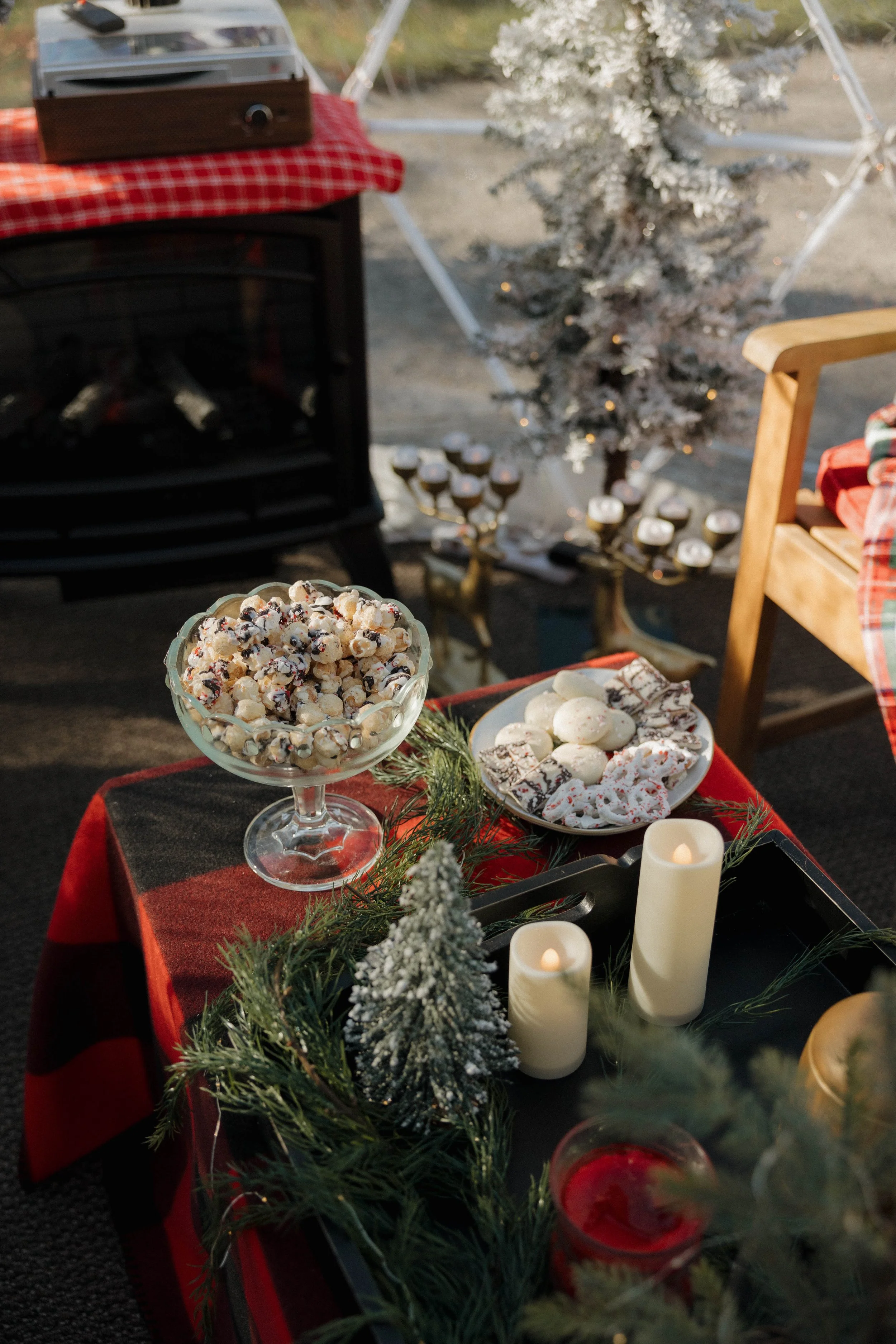 Table decorated with holiday treats, candles, and greenery in a cozy Christmas scene, with a small snow-dusted Christmas tree and a fireplace in the background.