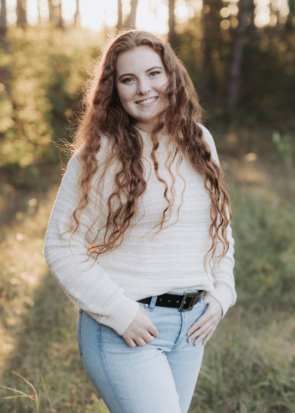 A young woman with long, curly red hair standing outdoors in a wooded area during golden hour, smiling at the camera.