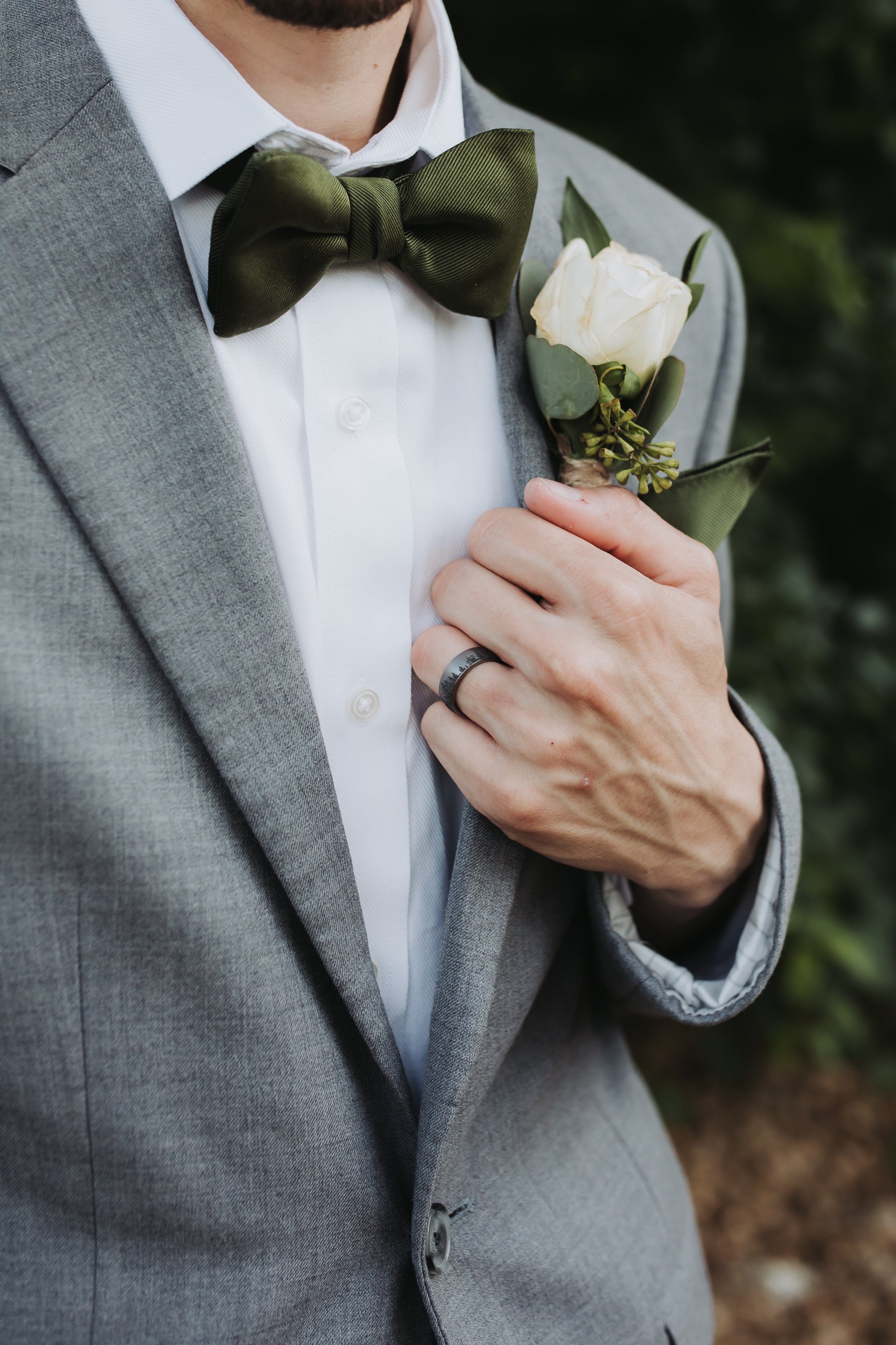 Close-up of a man wearing a grey suit and white shirt with a green bow tie, holding a white rose boutonniere on his chest, with a wedding band on his finger.