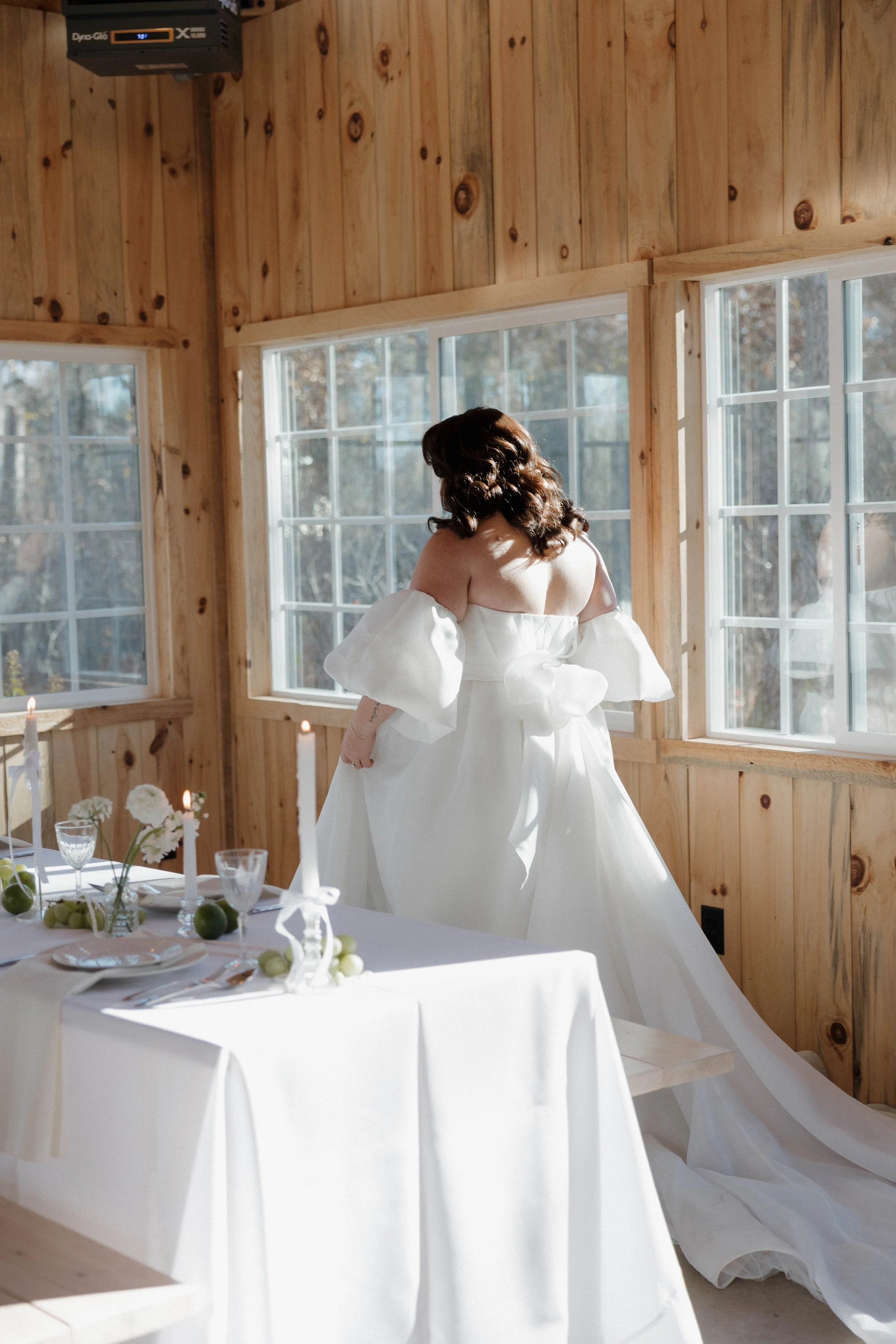 A bride in a white wedding dress with puffed sleeves stands near windows in a wooden-paneled room, with a table set for a celebration including candles, flowers, and glassware.