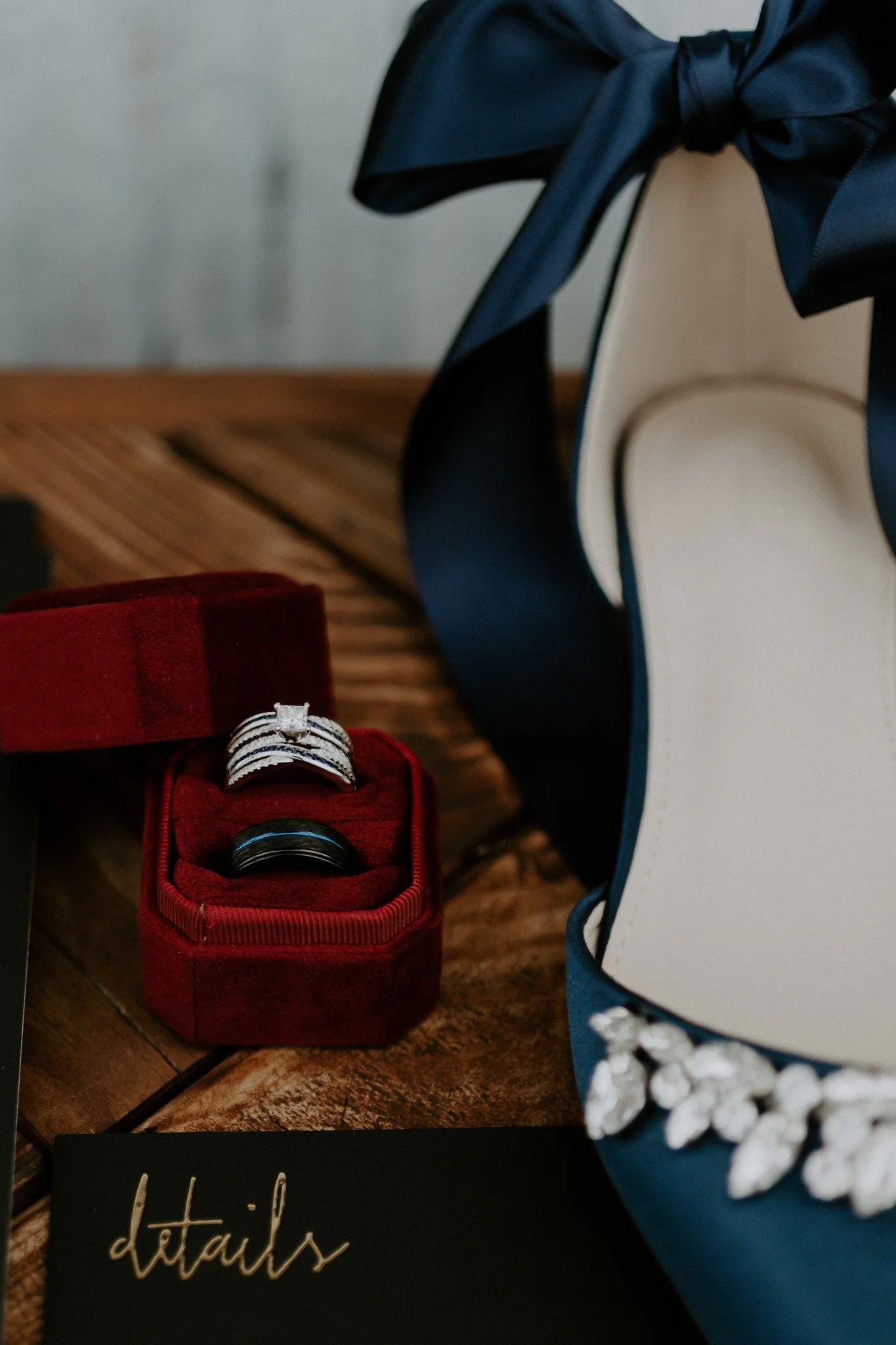 A close-up of a red ring box containing a diamond engagement ring, with black and silver wedding bands inside. A white purse with a navy blue ribbon and floral embellishment is partially visible on the right side. A black card with the word 'details'