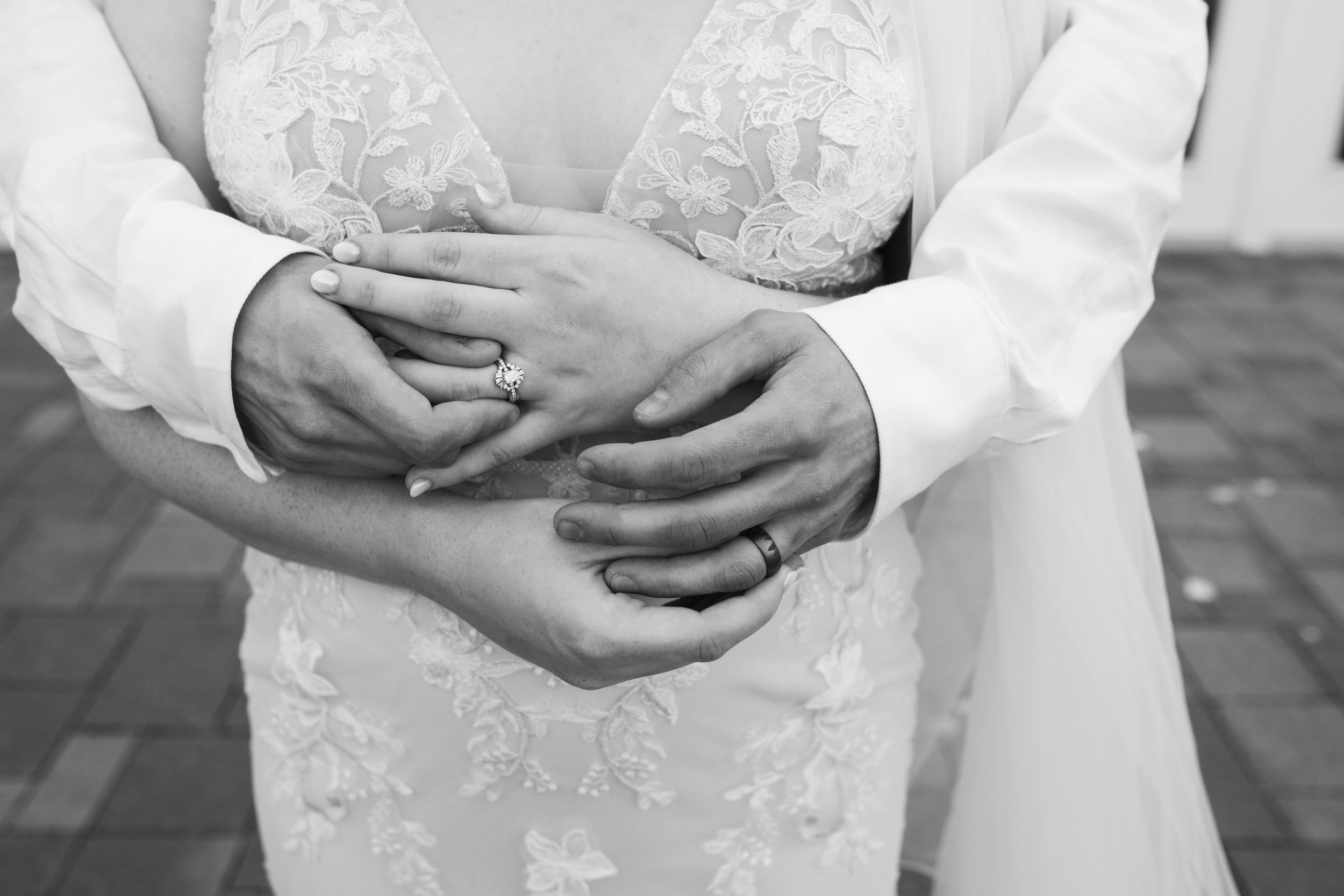 Black and white photo of a couple holding hands, showing wedding rings, with a lace wedding dress visible.