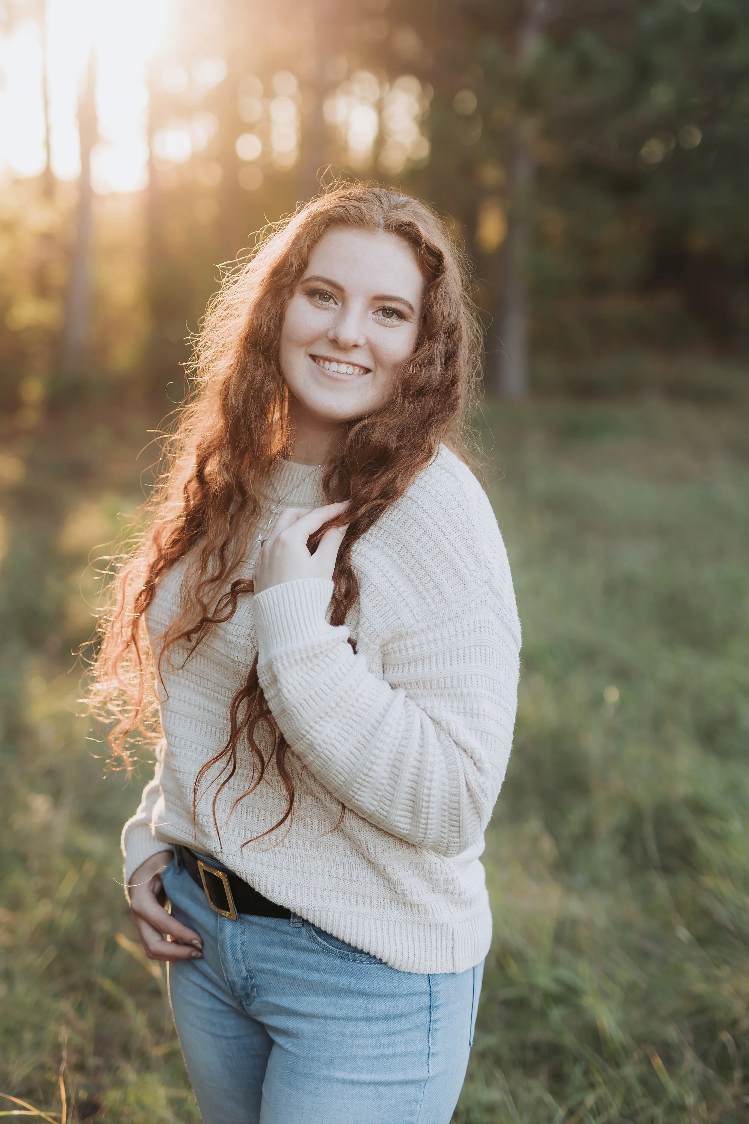 A young woman with long, curly red hair smiling outdoors during sunset, wearing a cream-colored sweater and light blue jeans.