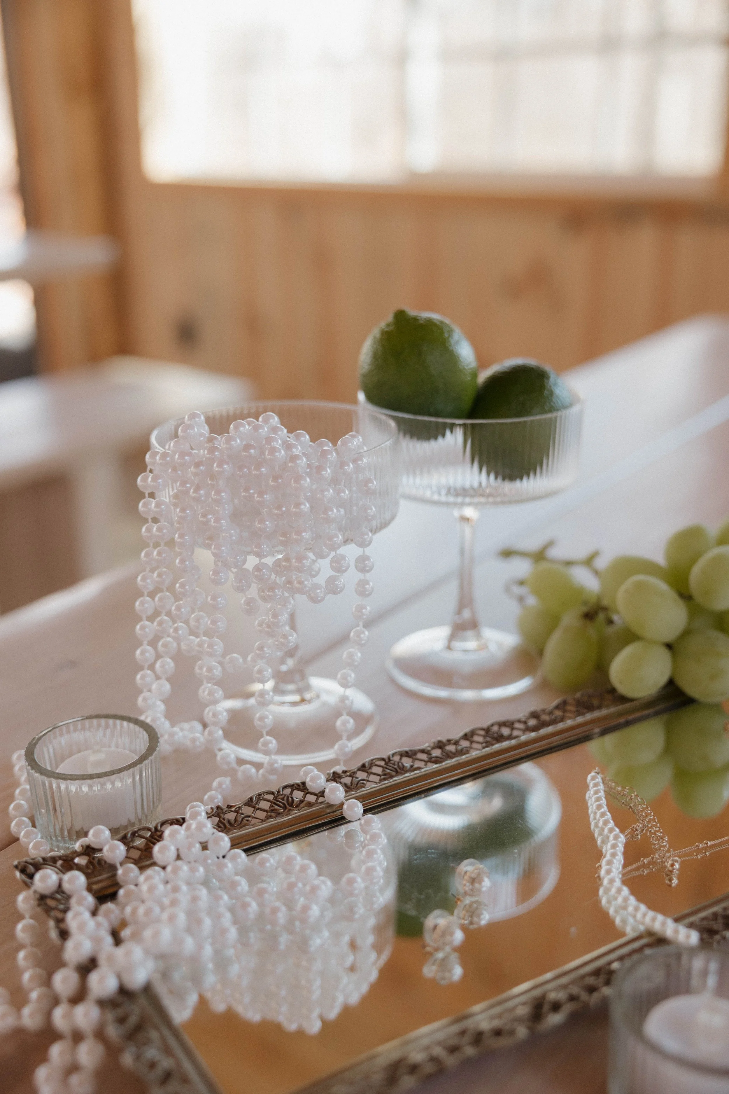 Decorative table setting with pearl necklaces, lime, and grapes, reflected on a glass surface.