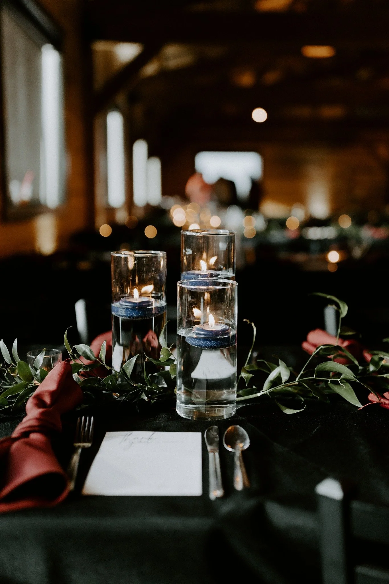 Lit candles in glass holders on a decorated table at a rustic indoor event.
