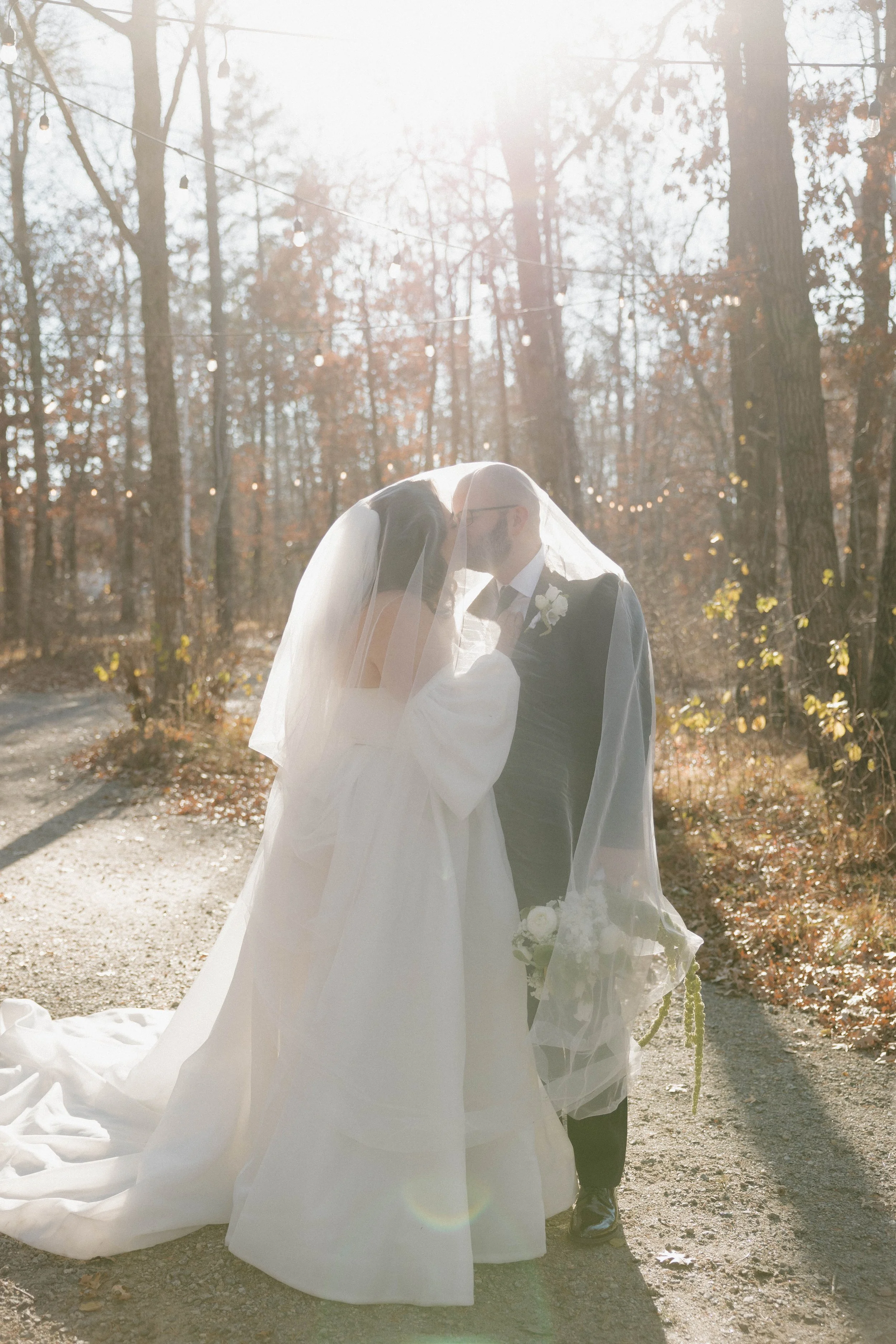 Bride and groom embracing outdoors in a wooded area, sunlight shining through the trees, wearing wedding attire, with the bride under a veil.