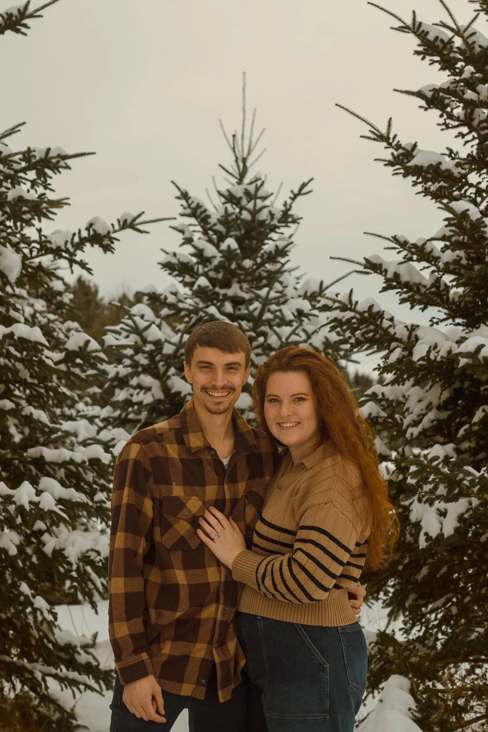 A smiling man and woman standing outdoors among snow-covered evergreen trees. The man has short brown hair and a beard, wearing a brown plaid jacket. The woman has long red hair, wearing a tan sweater with black stripes. They are embracing, with the woman's left hand showing an engagement ring.