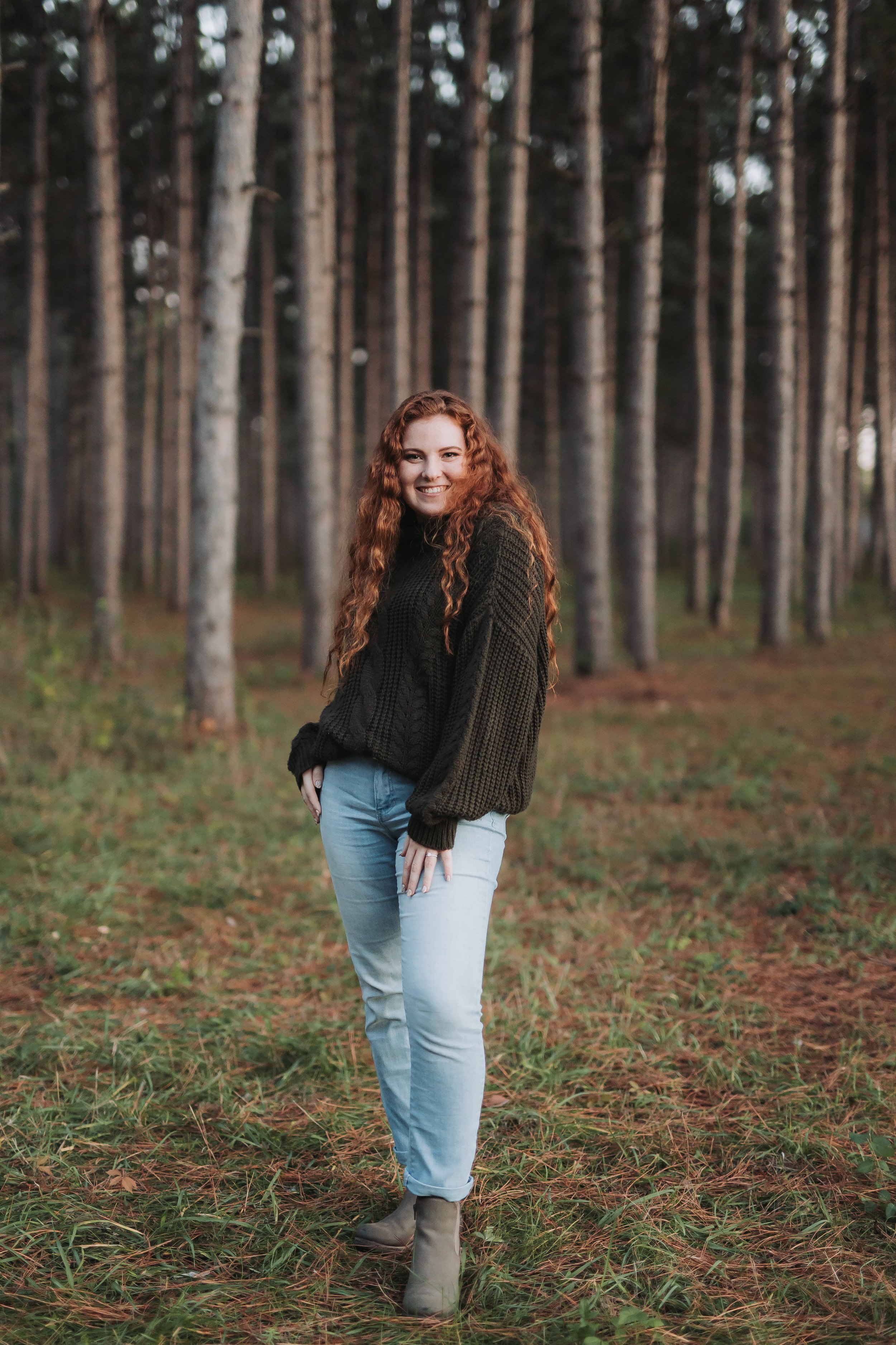 A woman with long curly red hair smiling and standing outdoors in a forest with tall trees in the background, wearing a dark sweater, light jeans, and ankle boots.
