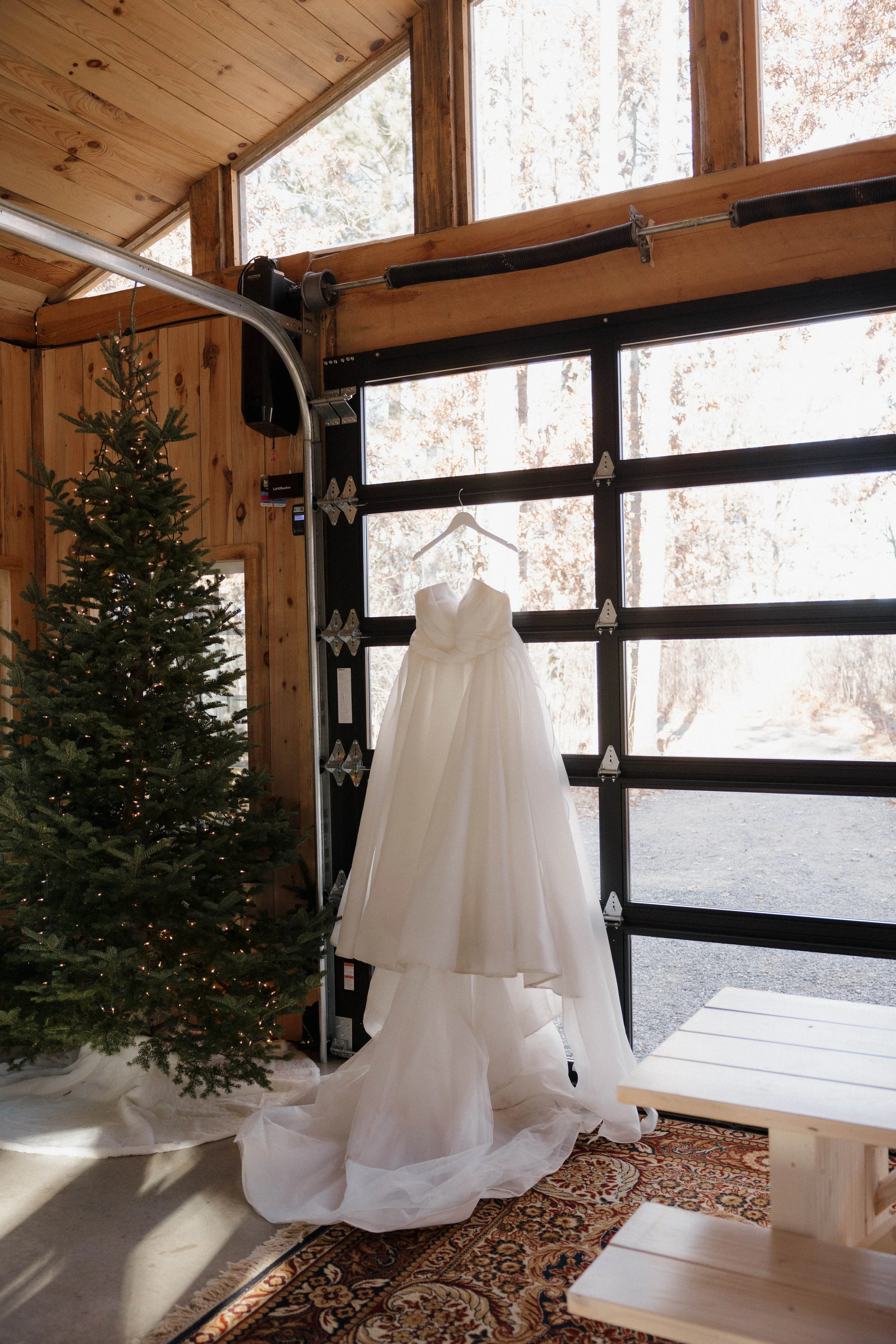 A white wedding dress hanging from a hanger on a garage door with a Christmas tree next to it inside a wood-paneled room with large windows.