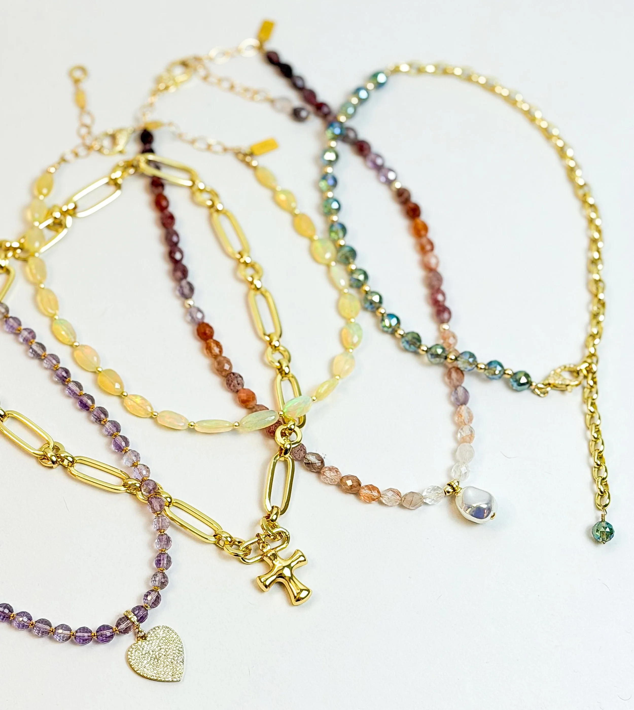 Multiple colorful necklaces with gemstone beads, chains, and pendants, including a gold crystal heart and a gold cross, displayed on a white background.