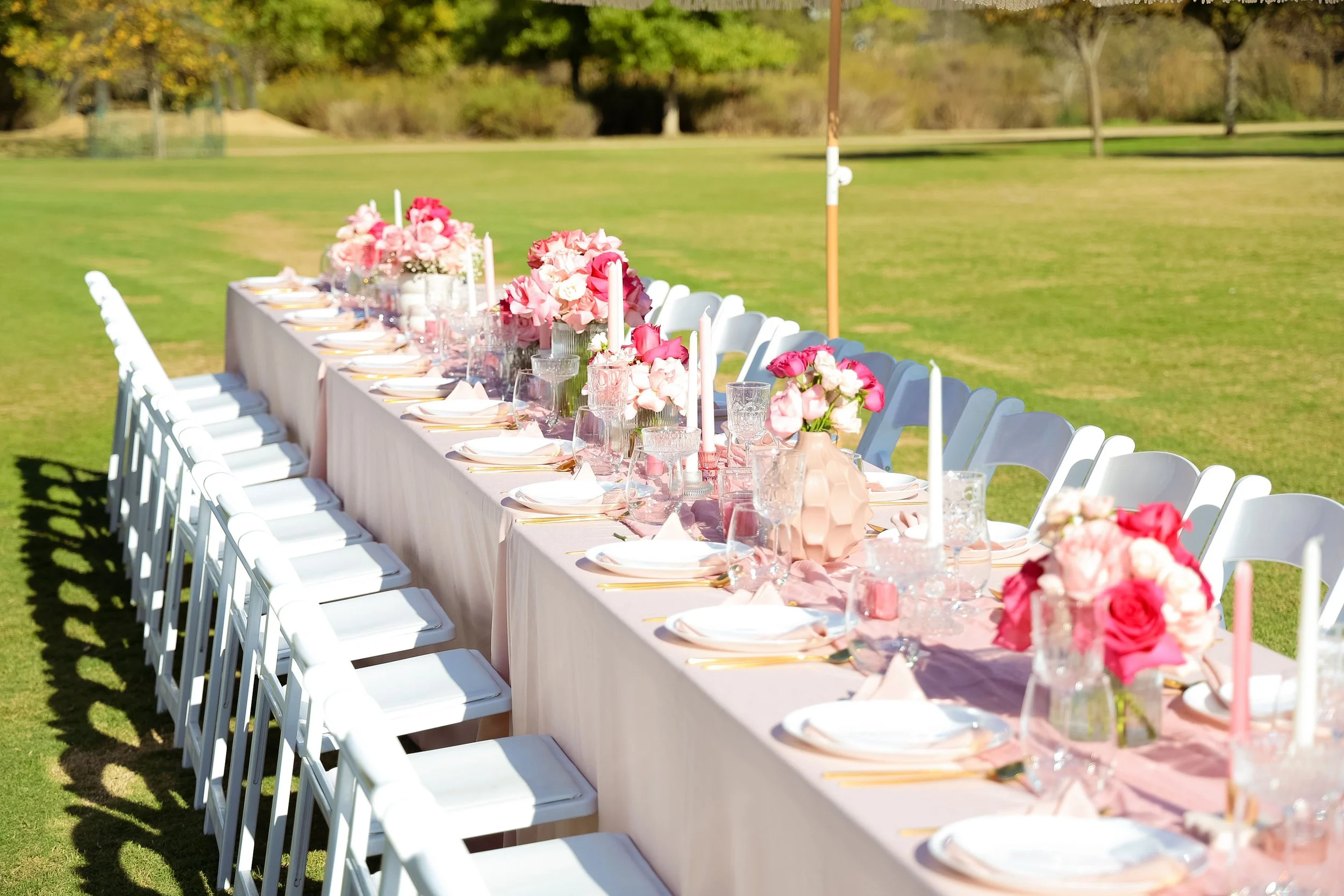 Long outdoor banquet table decorated with pink floral arrangements, candles, and glassware, set on a grassy field with trees in the background for a celebration or wedding.