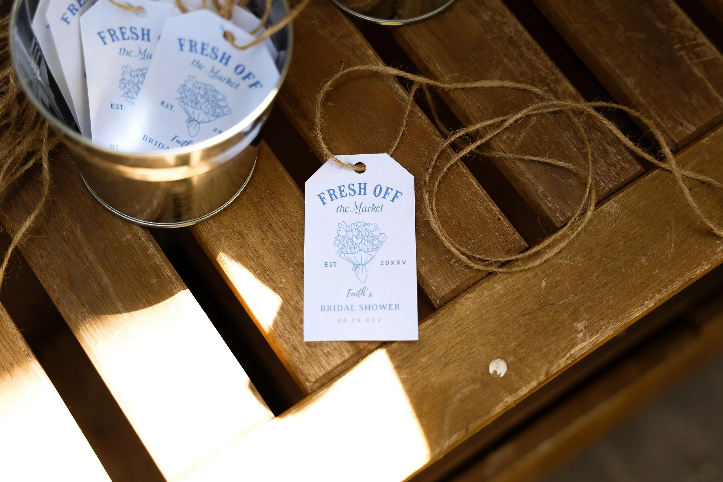 A metal container filled with paper tags that read 'FRESH OFF the Market,' with a floral illustration, date, and event details for a bridal shower, placed on a wooden surface with a piece of rope nearby.