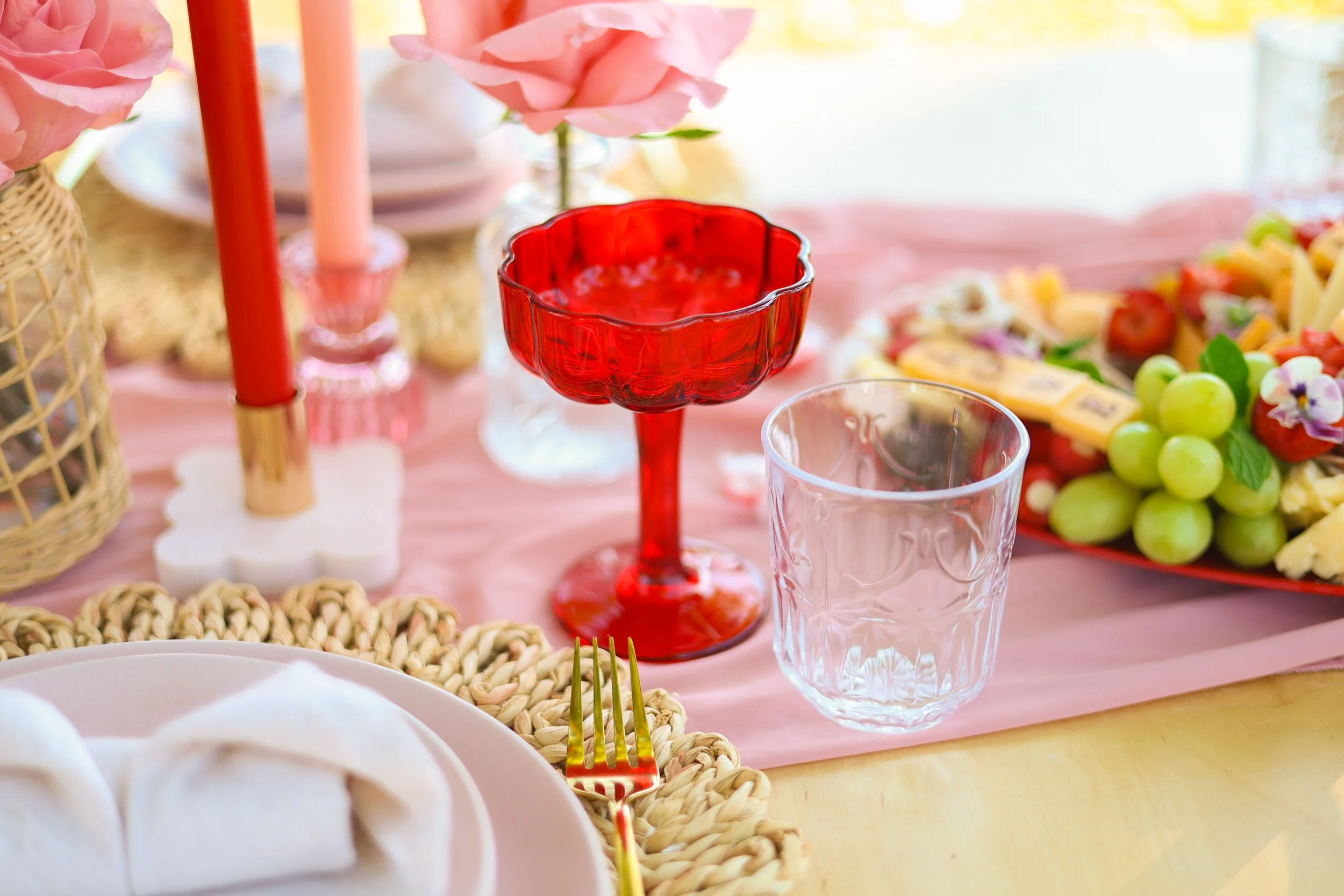 A table setting with pink tablecloth, a red glass goblet, a clear glass, a plate with napkin, gold fork, pink candles, a centerpiece of flowers (pink roses), and a colorful fruit platter with grapes and other fruits.